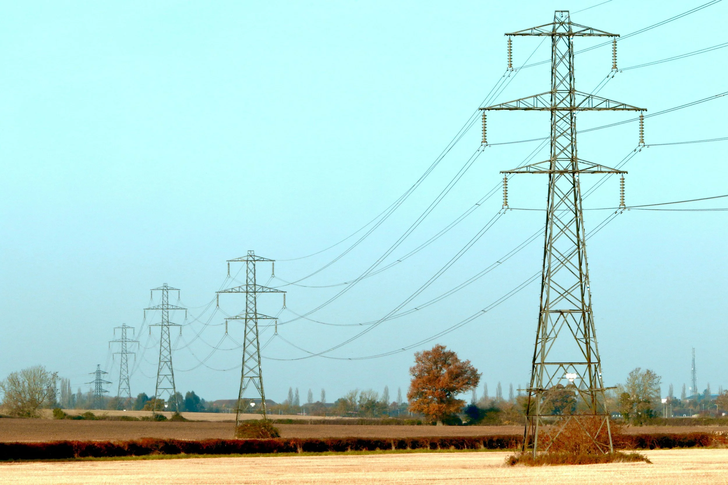 A series of high-voltage power line towers extending into the distance across a rural landscape with trees and fields, under a clear blue sky.