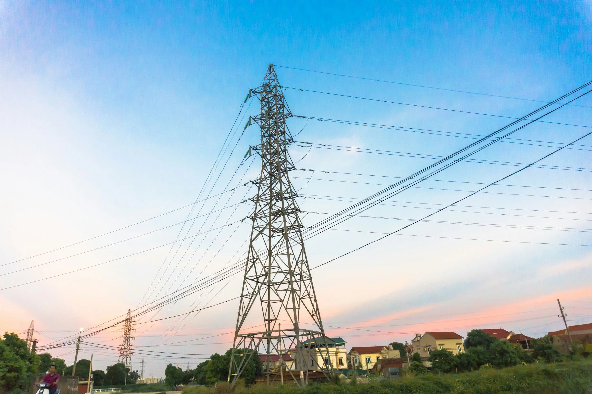 An electricity transmission tower with power lines during sunset, with houses and trees in the background.
