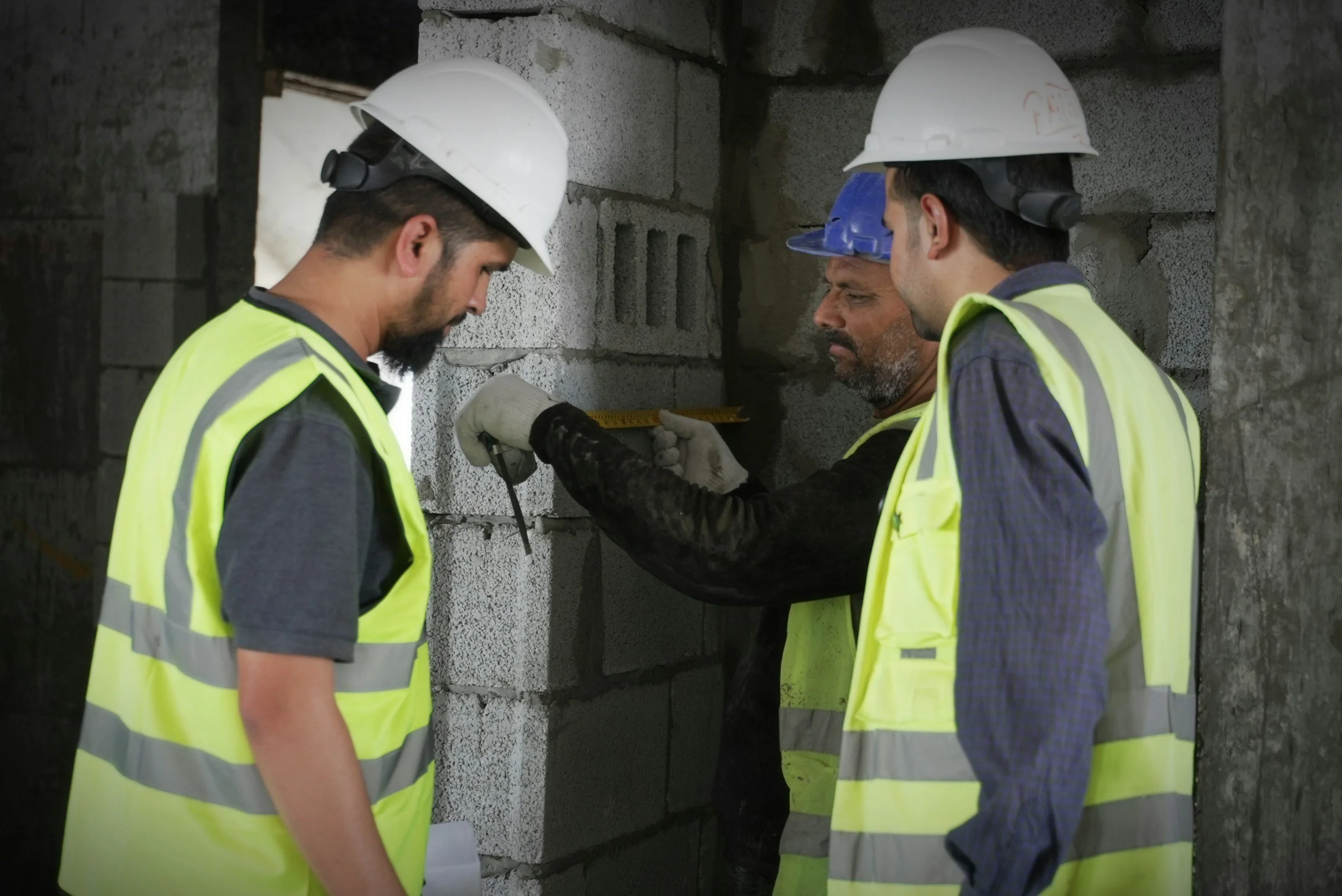 Three construction workers wearing helmets and safety vests inspecting a brick wall at a construction site.
