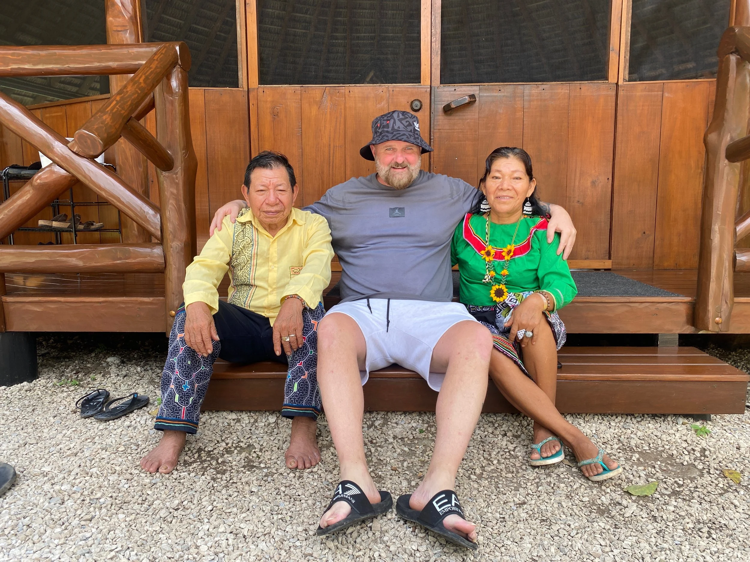 Three people sitting on a wooden platform in front of a wooden building. Two women wearing colorful traditional outfits and a man in casual shorts and t-shirt with a bucket hat.