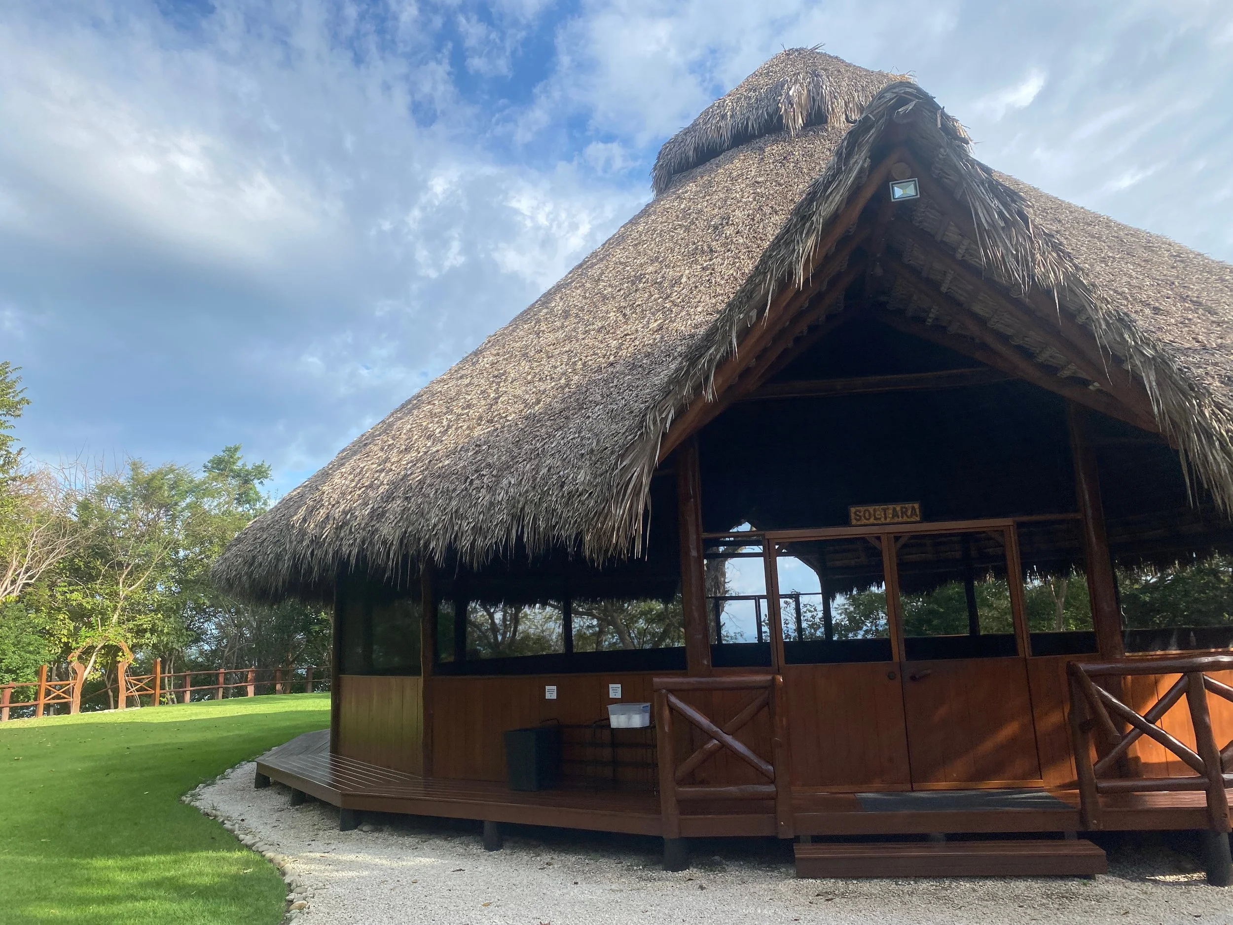 A wooden hut with a thatched roof, situated on a grassy area with trees in the background, under a partly cloudy sky.
