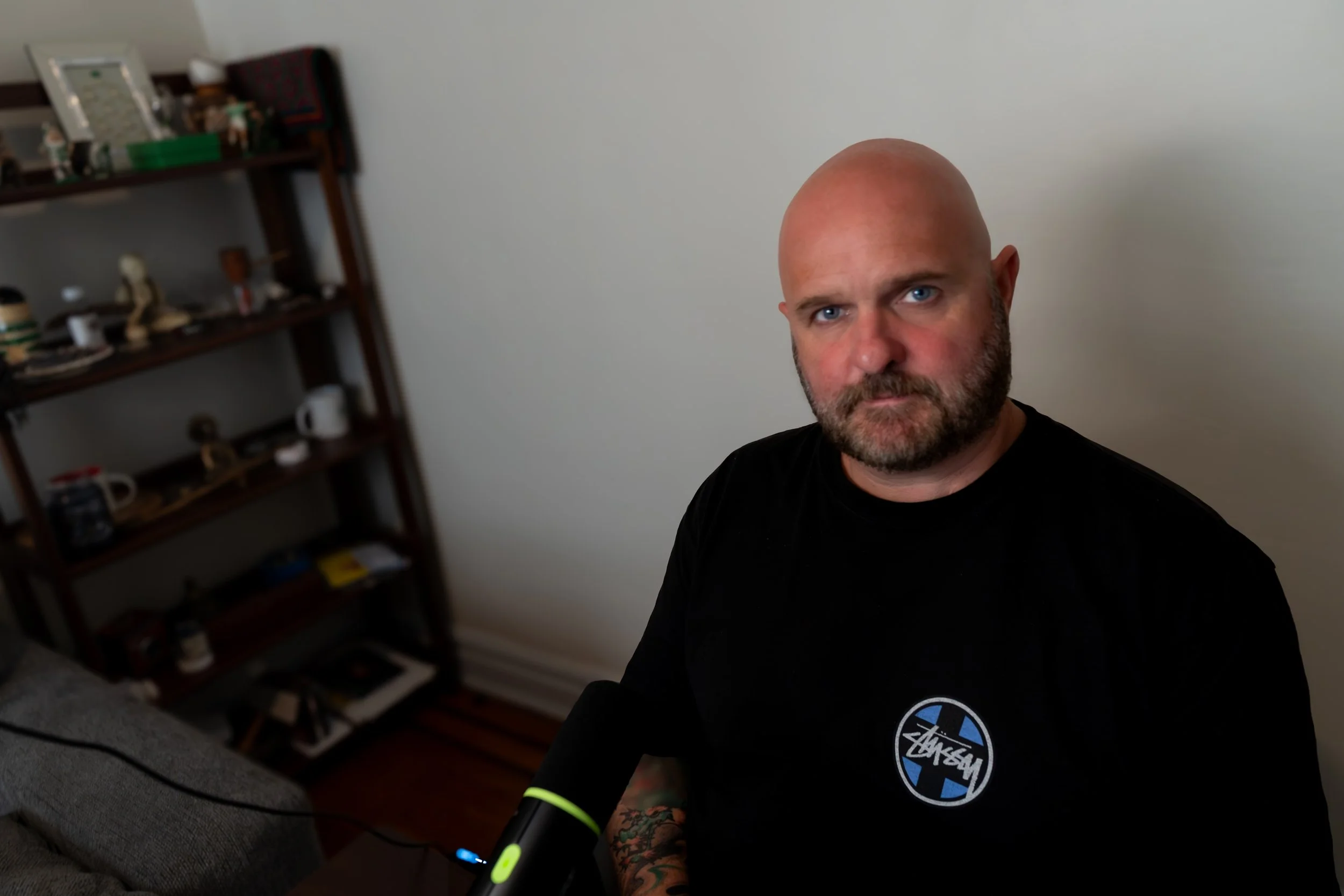 A bald man with a beard and blue eyes sitting indoors, wearing a black t-shirt with a logo on it, in front of a white wall. A gray sofa and a wooden shelf with various decorative items and mugs are visible in the background.