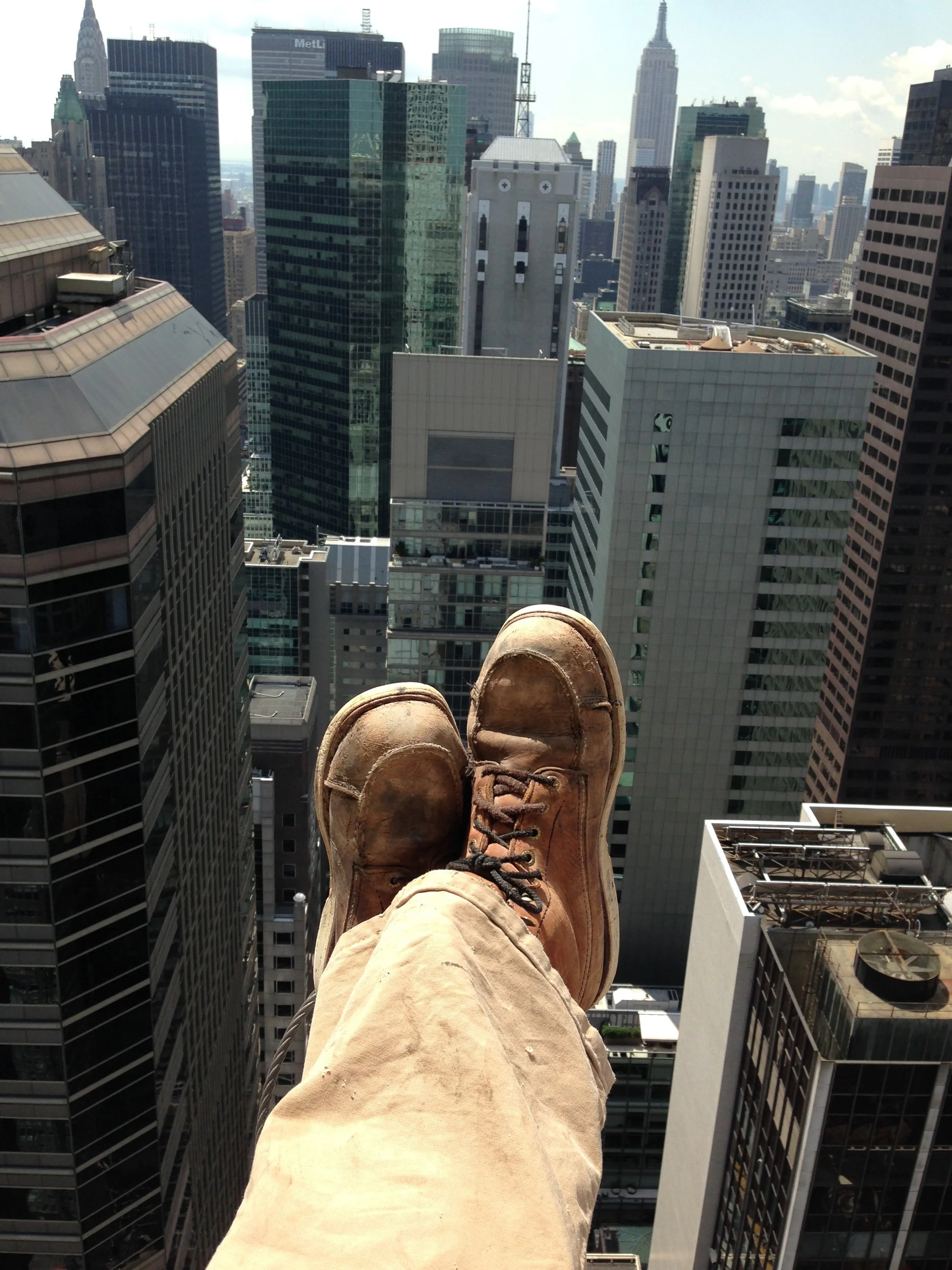 Person standing on a rooftop overlooking a cityscape with tall skyscrapers, including the Empire State Building, wearing tan pants and brown leather boots.