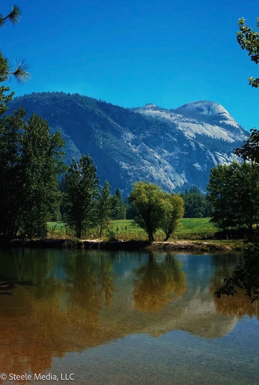 A scenic landscape with a mountain in the background, trees along a river or lake in the foreground, and a clear blue sky.