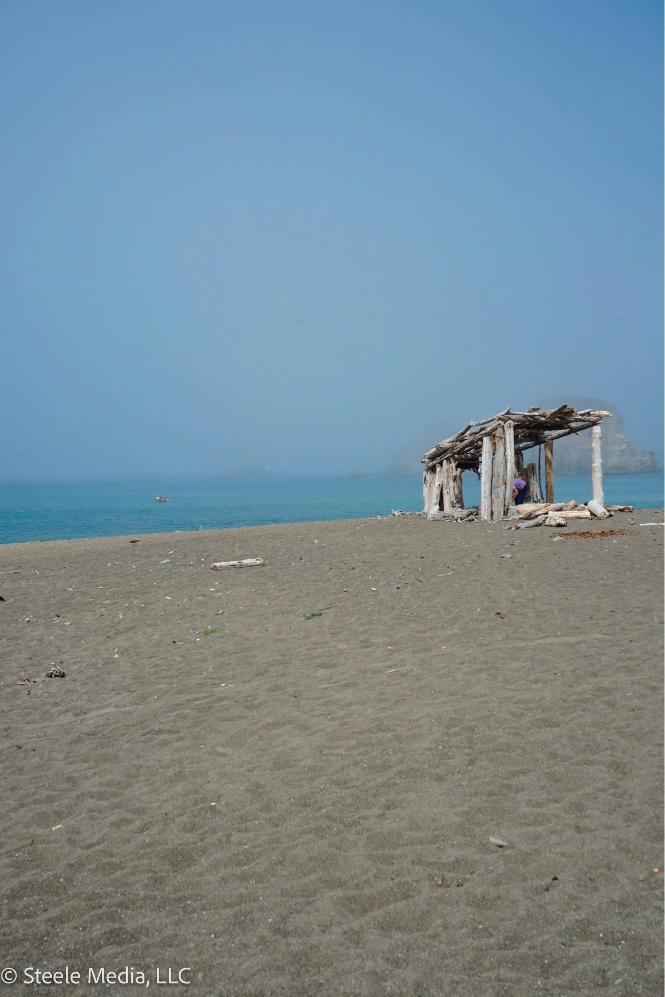 A sandy beach with a simple wooden structure near the shoreline under a light blue sky, with a distant rocky island and a boat in the water.