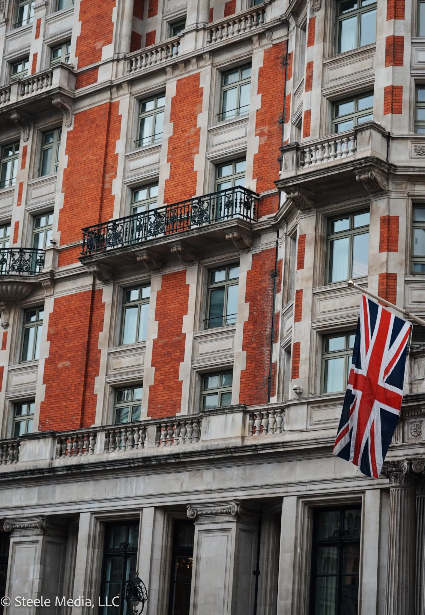 Close-up of a building with red brick and white stone architecture, featuring windows with decorative frames and small balconies with black wrought iron railings, and a Union Jack flag hanging outside.