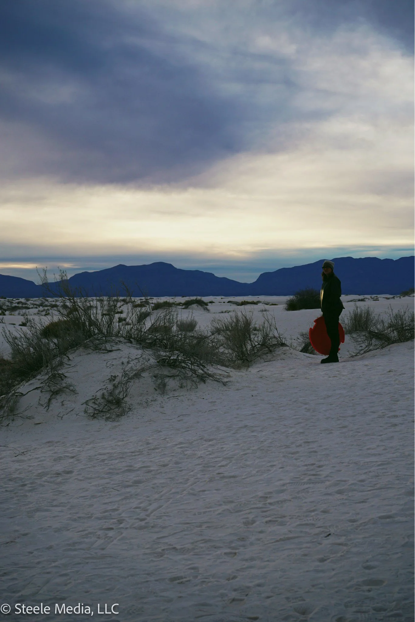 A person standing on a sandy desert landscape holding a red sled, with mountains in the background and a cloudy sky overhead.