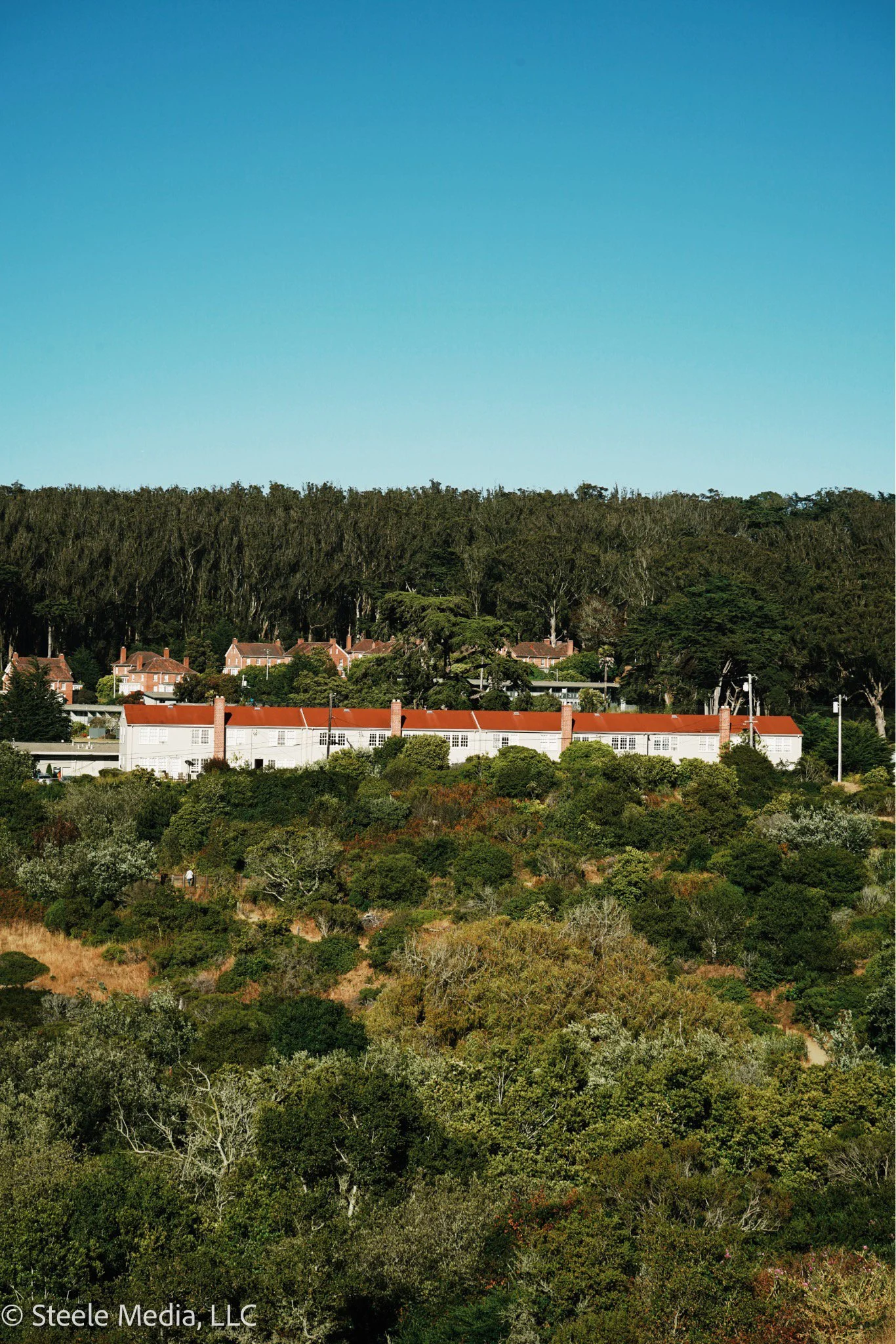 A landscape with dense trees in the foreground, a white building with a red roof in the middle, and a forested hillside under a blue sky in the background.