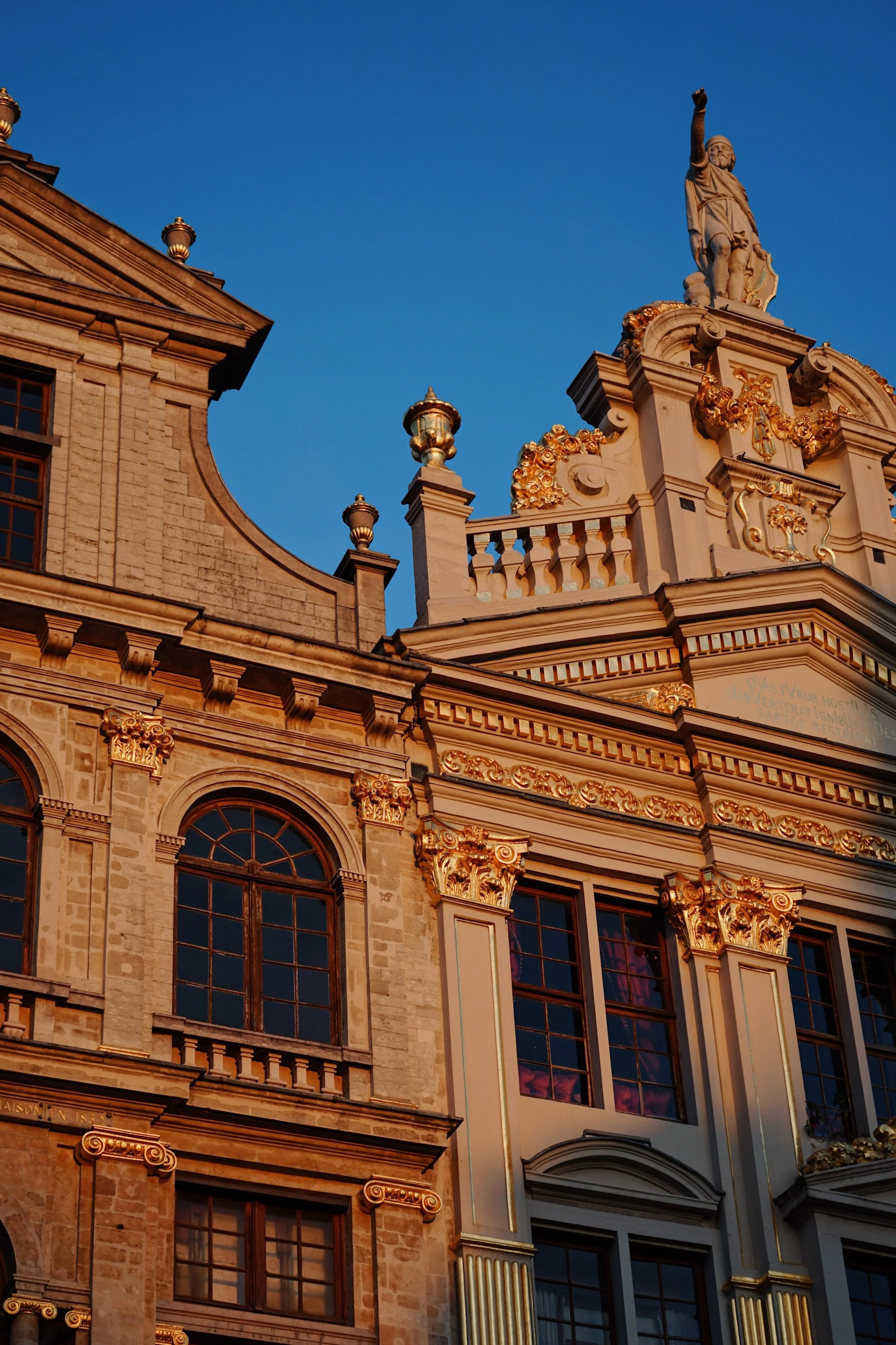 Close-up of the ornate facade of a historic building with gold accents, large arched windows, and a statue on top against a clear blue sky.