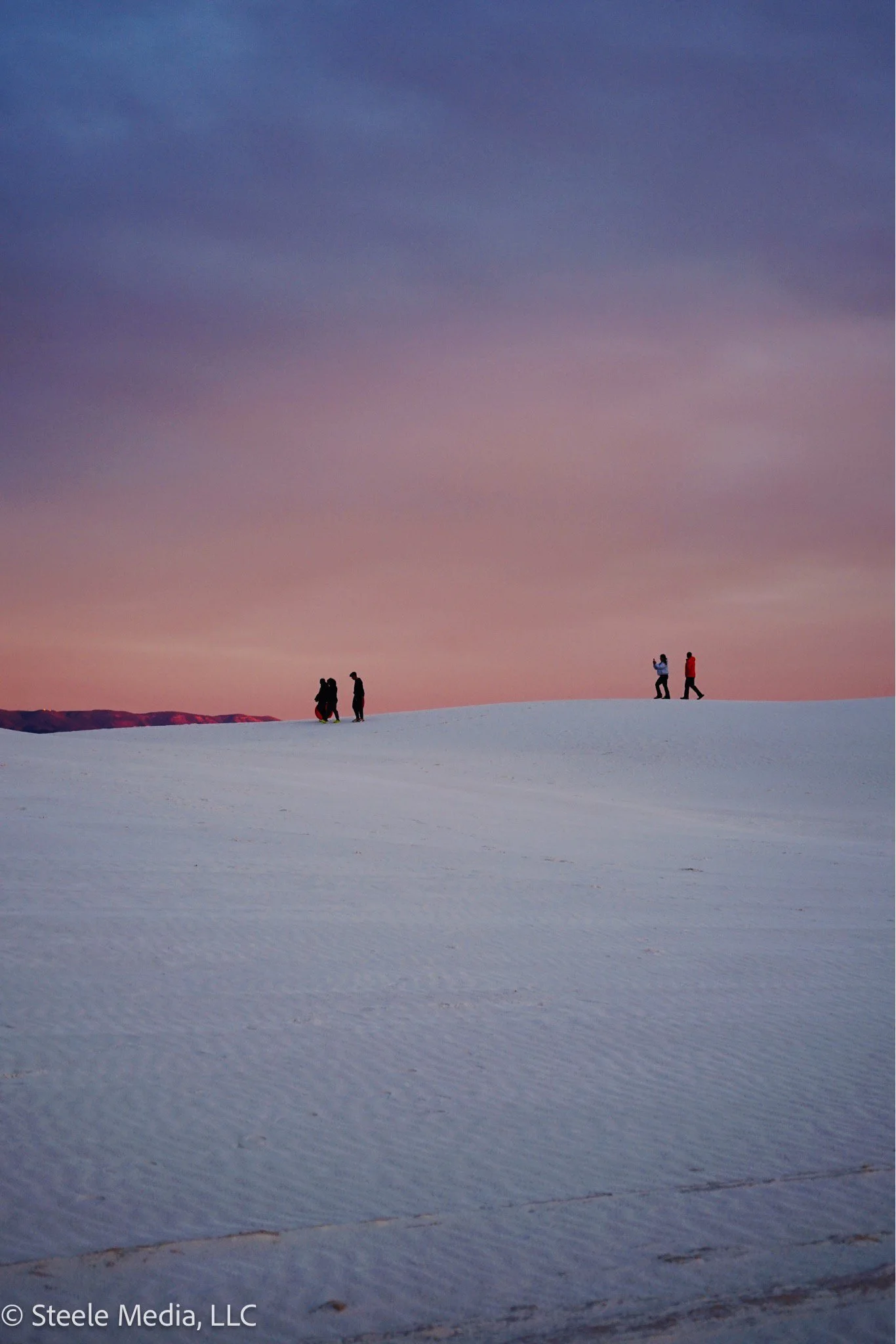 Silhouettes of six people on a snow-covered landscape during sunset or sunrise with a colorful sky.