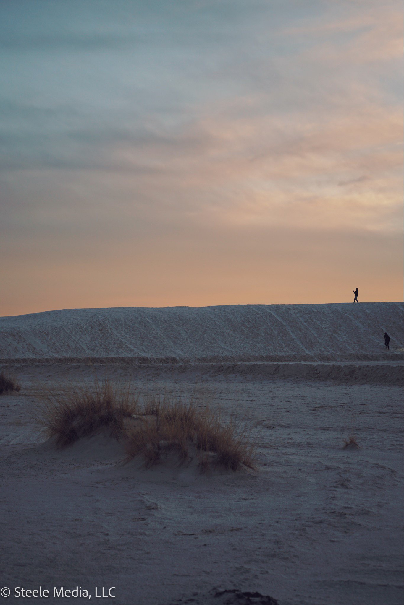 A landscape scene at sunset with a sandy, dune-filled area and sparse vegetation in the foreground. Two people are walking along the crest of a large dune, one near the top and one lower down, against a sky with soft, colorful clouds.