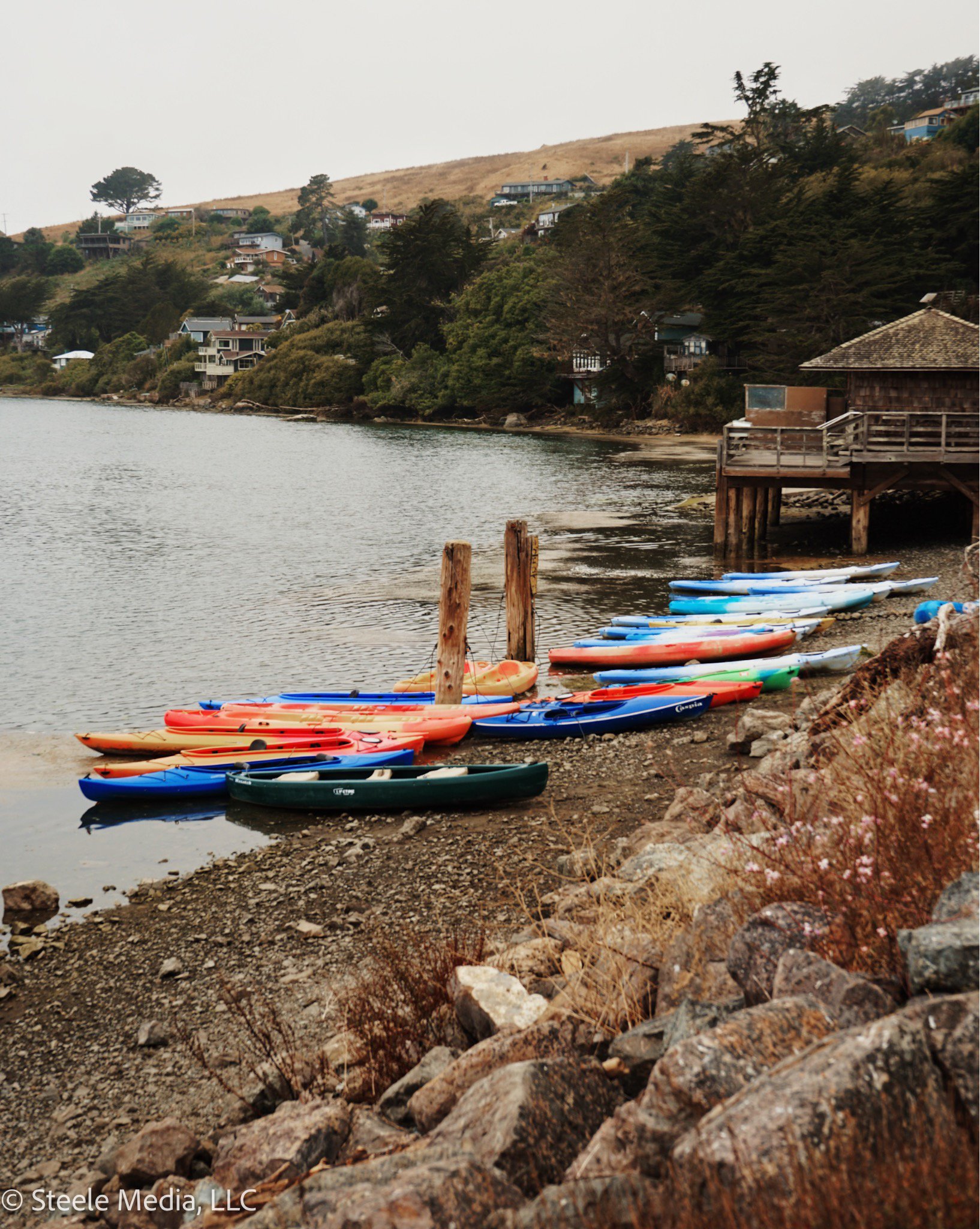 Row of colorful kayaks lined up on a rocky shoreline next to a body of water, with houses and trees on a hillside in the background.