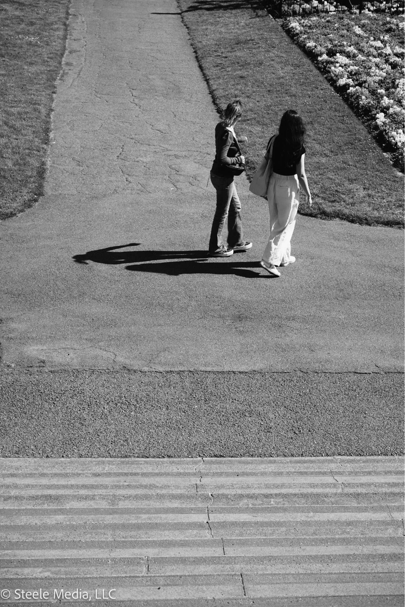 Black and white photo of two women walking on a sidewalk, with their long shadows cast on the ground. One woman is wearing a dark top and light pants, holding a bag, while the other woman is in a dark top and wide light pants, carrying a bag over her