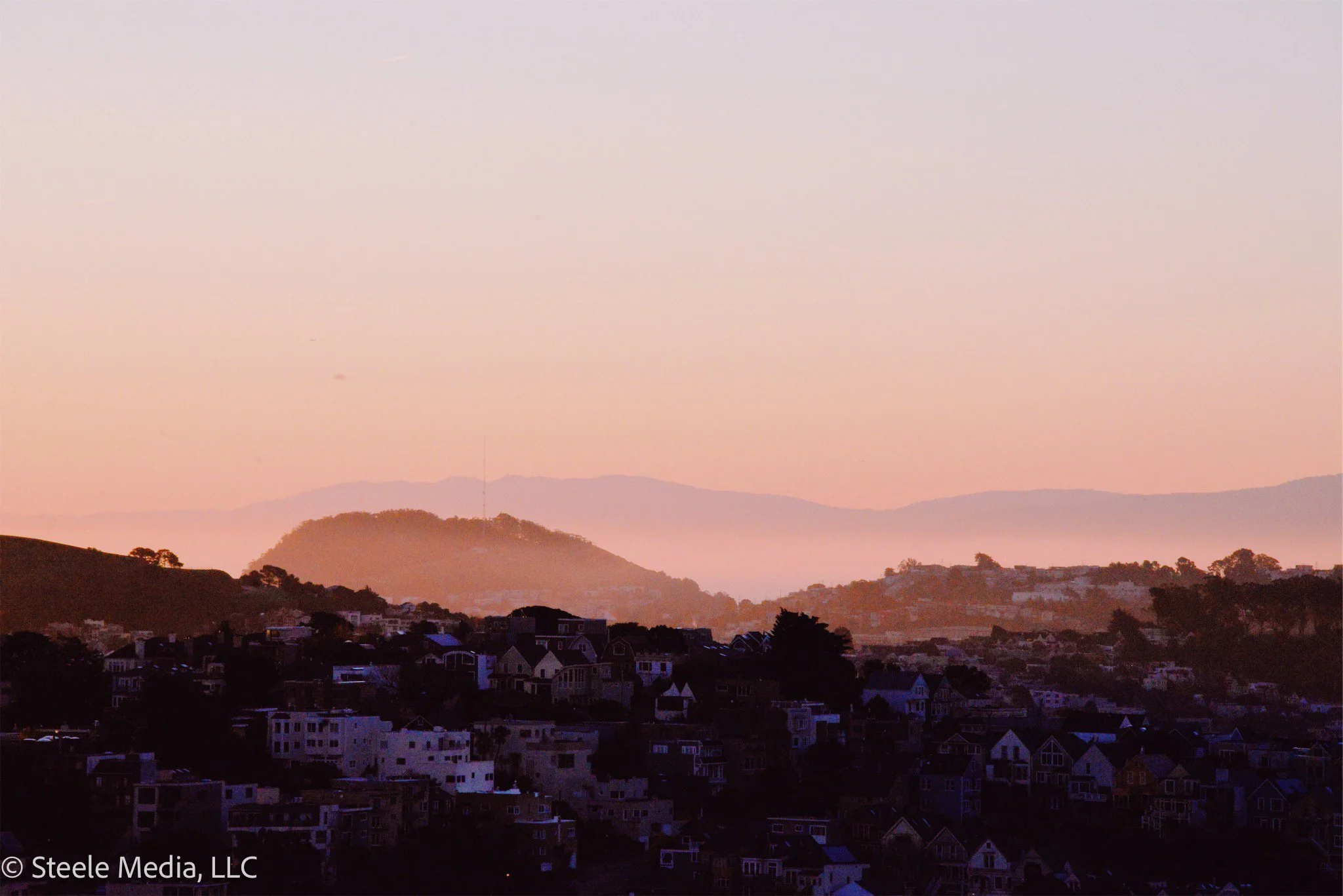 Hilly landscape at sunset with a cityscape in the foreground and mountains in the background, pink and purple sky.