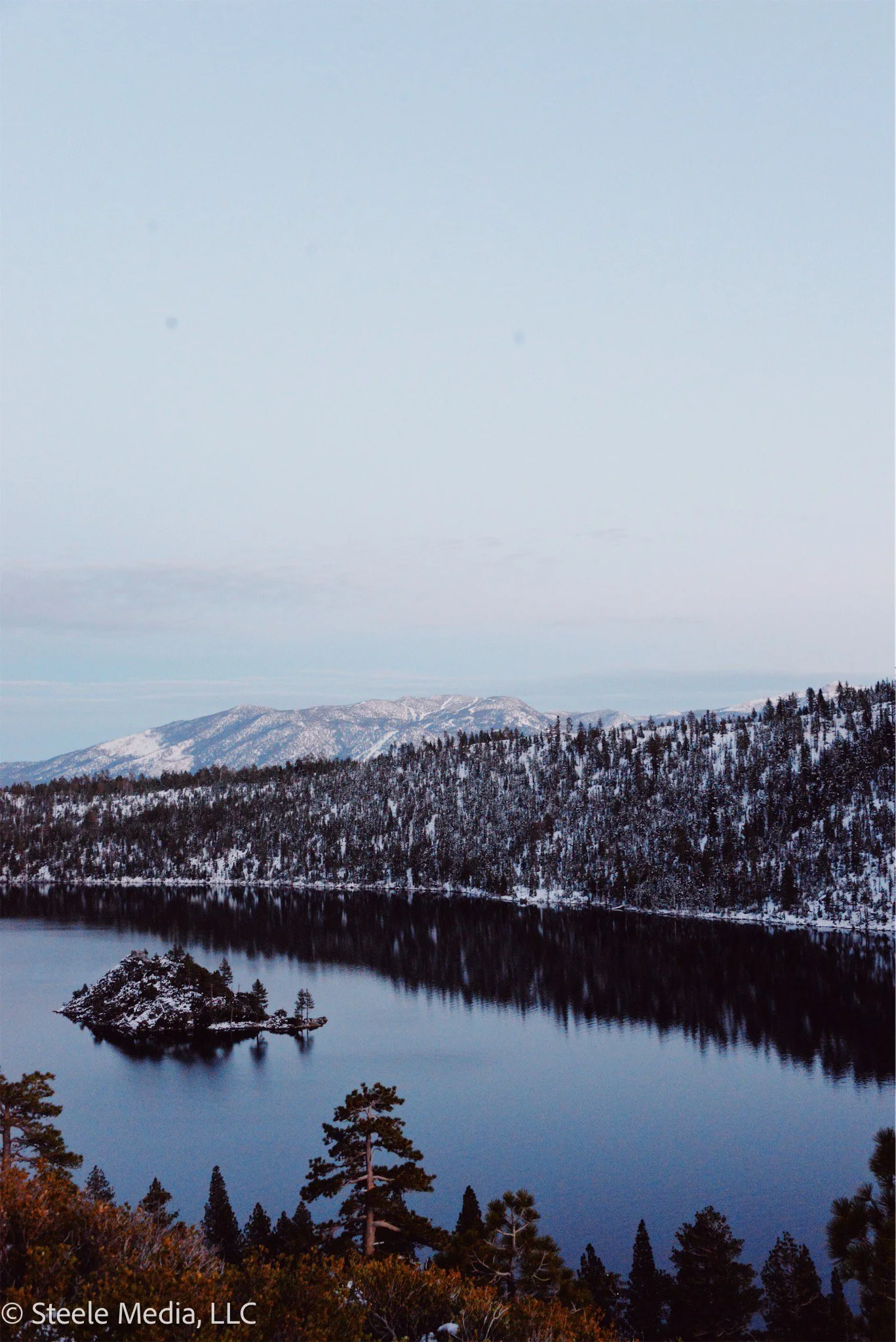Snow-covered mountains and trees reflected in a calm lake under a cloudy sky.