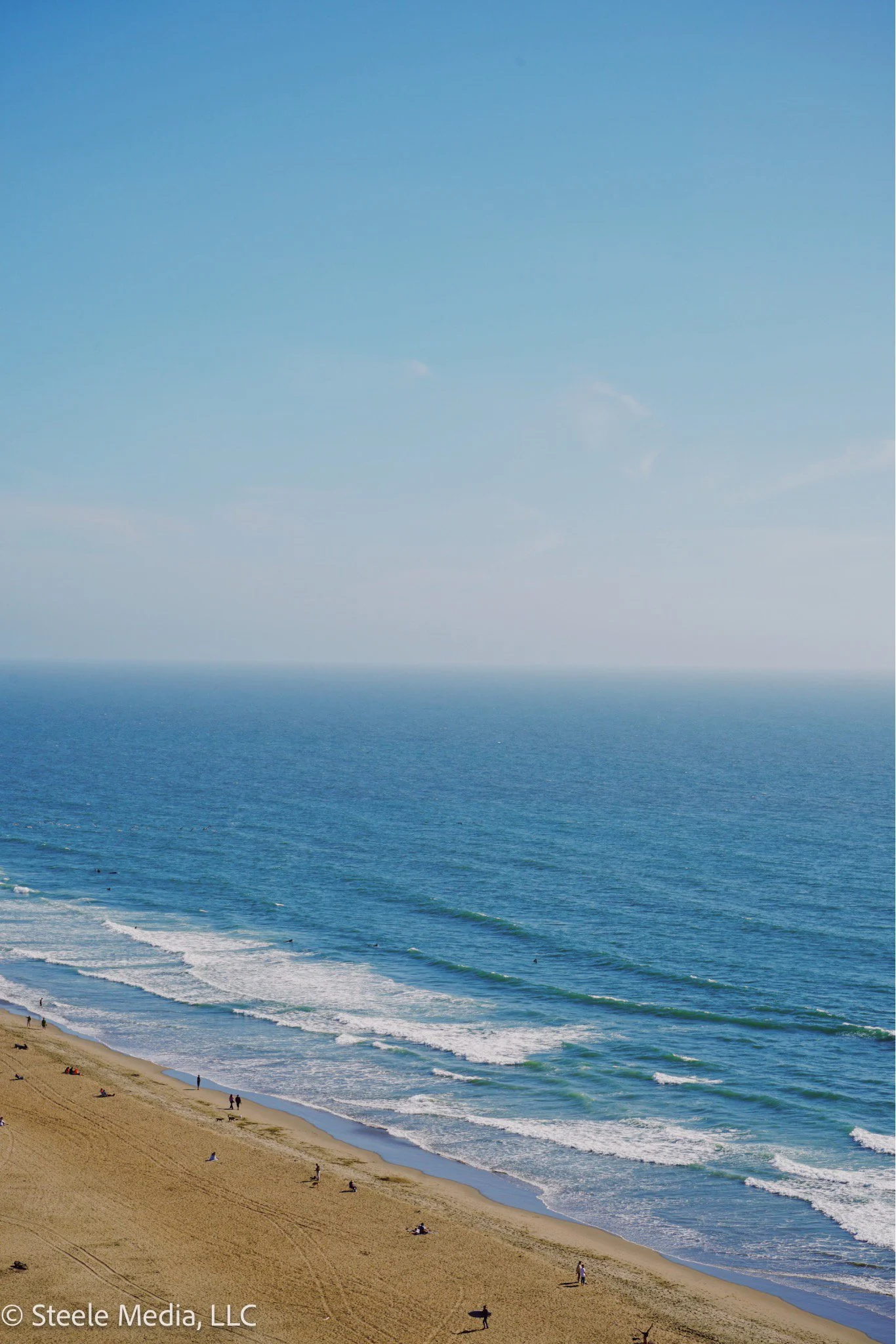 A view of a sandy beach with people walking and relaxing, along with ocean waves and a blue sky.