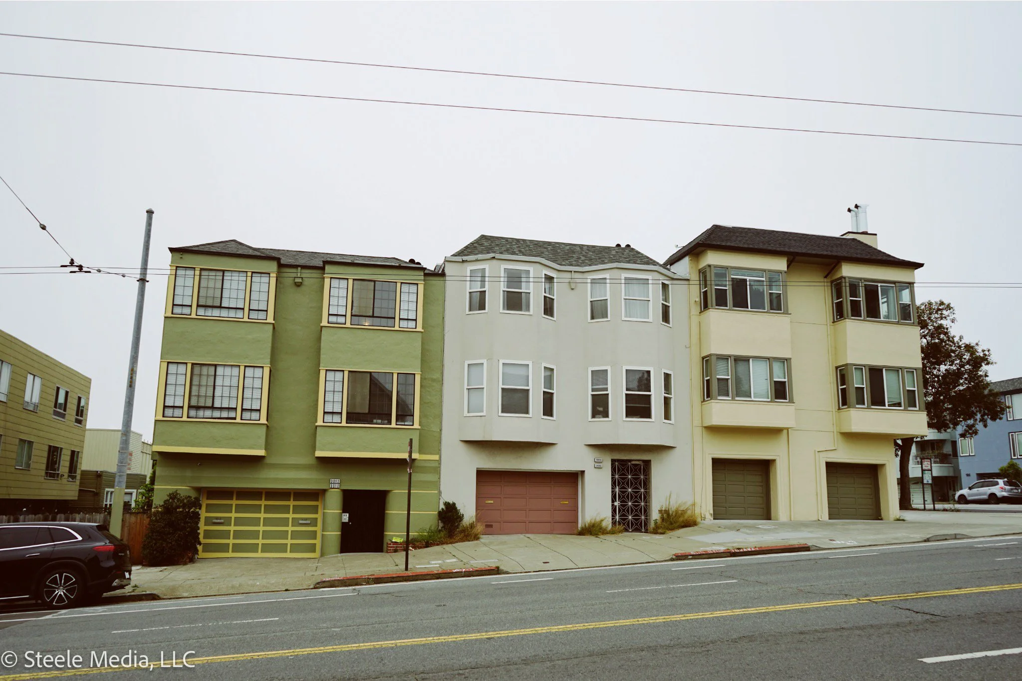 Three adjacent multistory residential buildings with garages on the ground level. The left building is green with a yellow garage door, the middle is light beige with a brown garage door, and the right building is pale yellow with two dark green gara