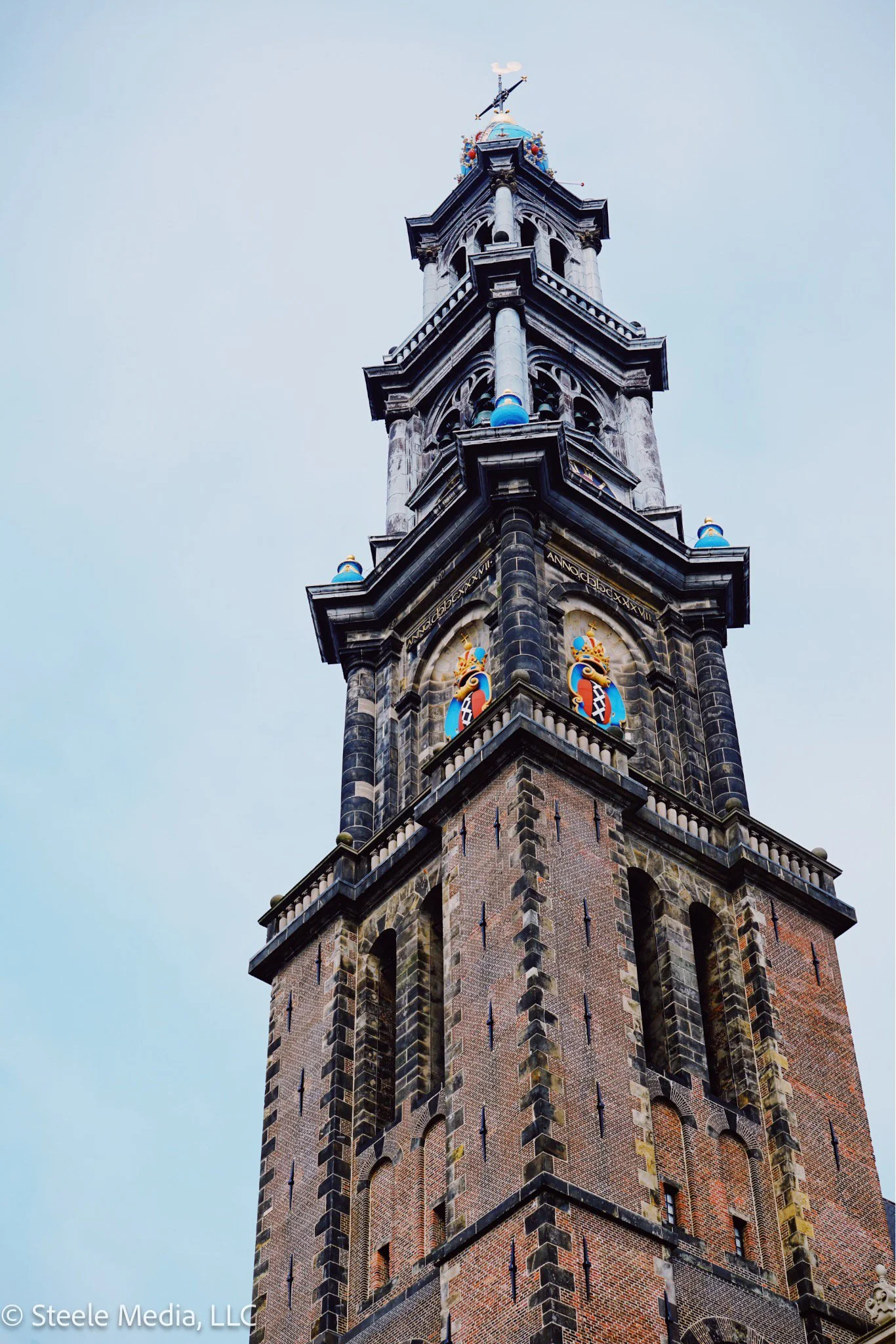 Close-up of a tall, historic clock tower made of brick and stone, with colorful decorative features near the clock face, and a weather vane at the top against a cloudy sky.