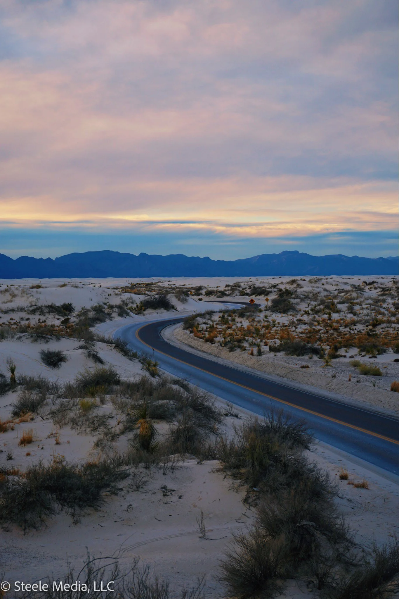 A winding road through a sandy desert landscape with sparse desert plants, mountains in the distance, and a colorful sky with pink and purple clouds.