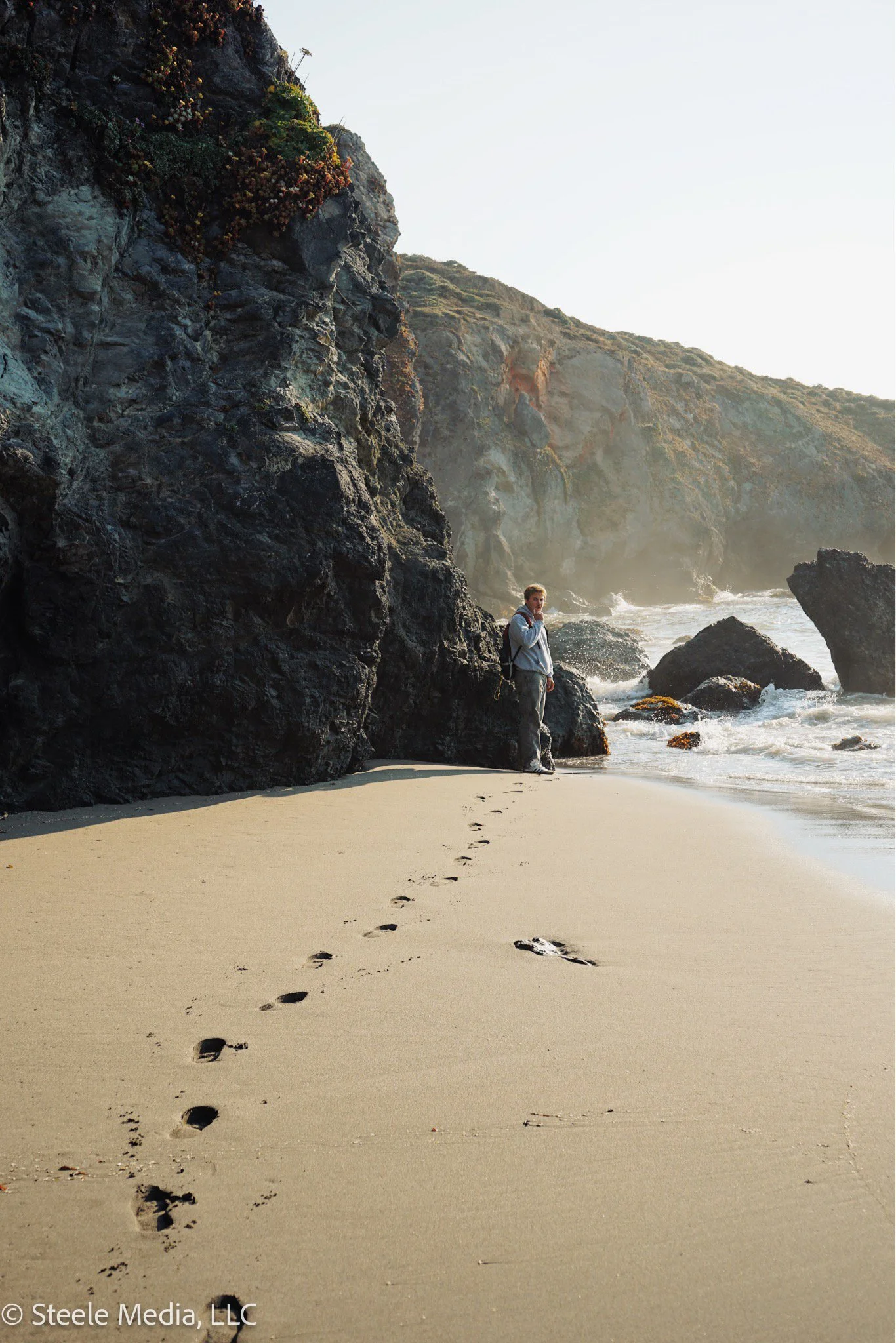 A person standing on a sandy beach near rocky cliffs, with footprints leading to them, ocean waves crashing against the rocks, and mist in the background