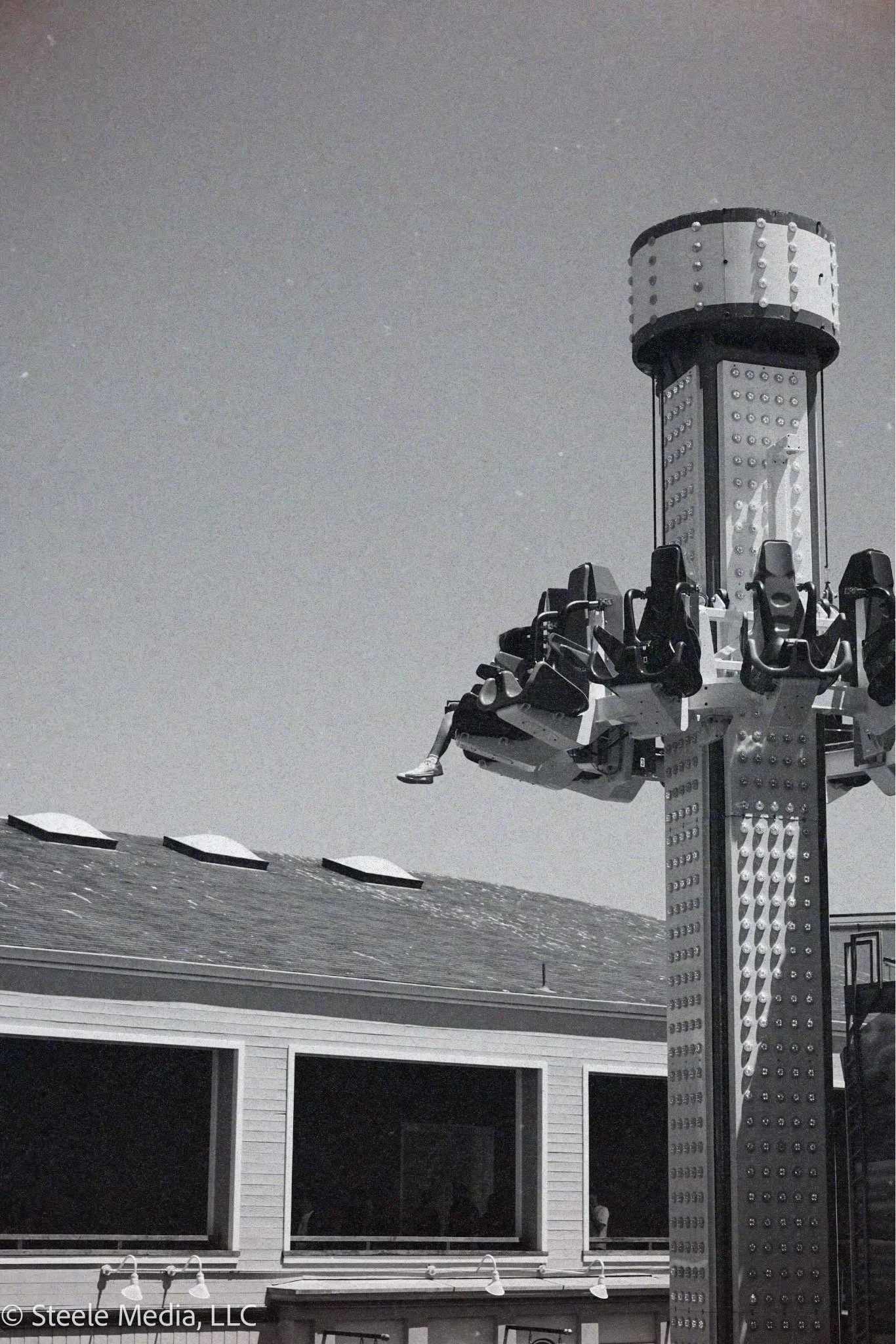 A black and white photo of a carnival ride with several empty seats hanging from a tall central pole, set against a night sky and building with windows in the background.