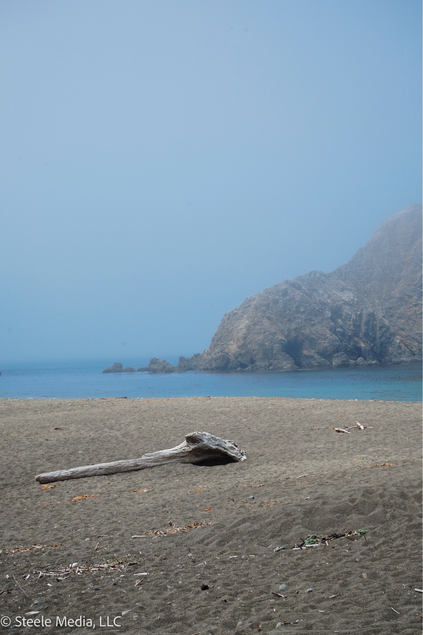 A sandy beach with driftwood and small debris, calm blue water, and large rocky cliffs under a hazy sky.