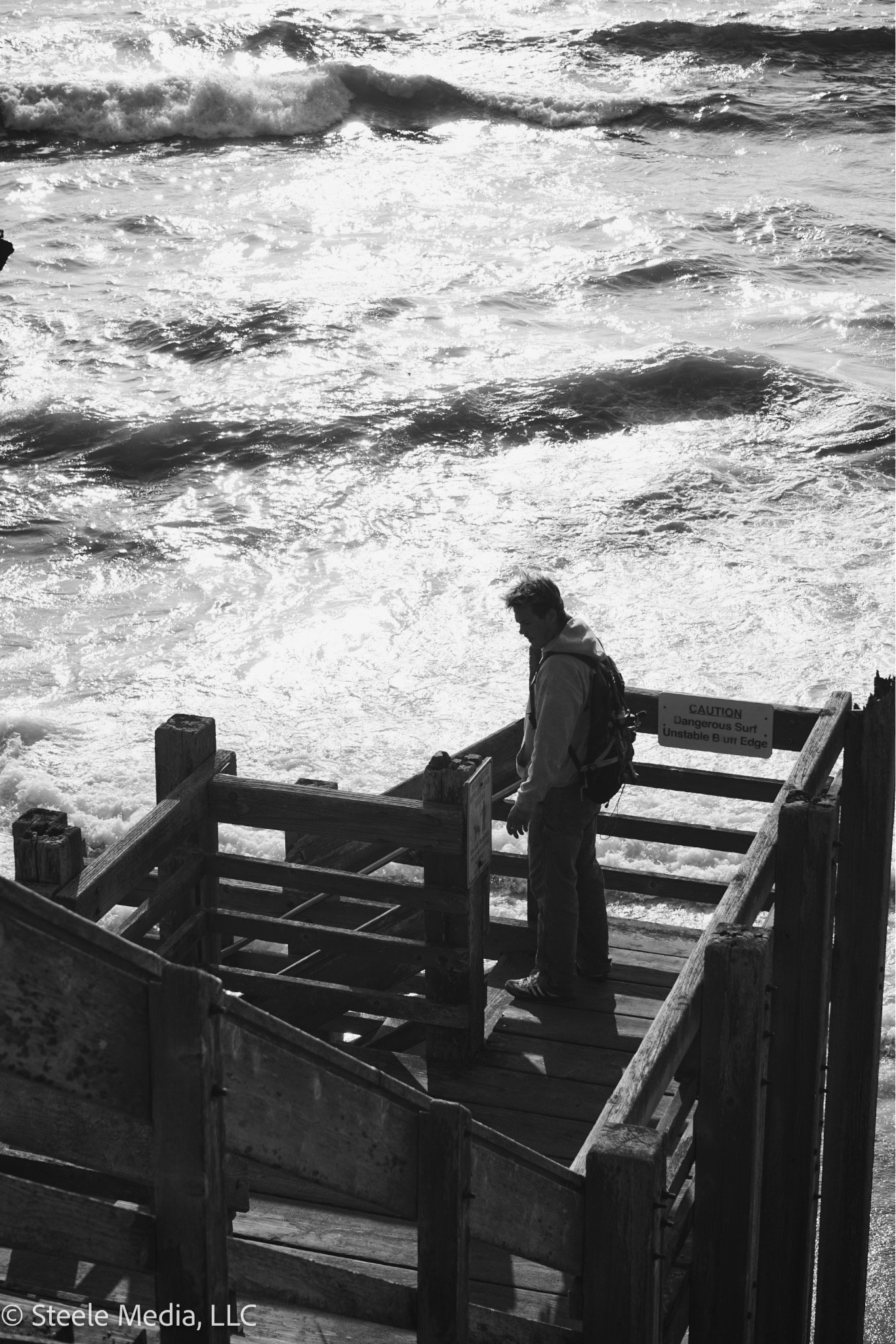 A person stands on a wooden pier near the ocean, looking down, with waves crashing behind them. The photo is in black and white, and there is a caution sign warning about dangerous surf and unstable edge.
