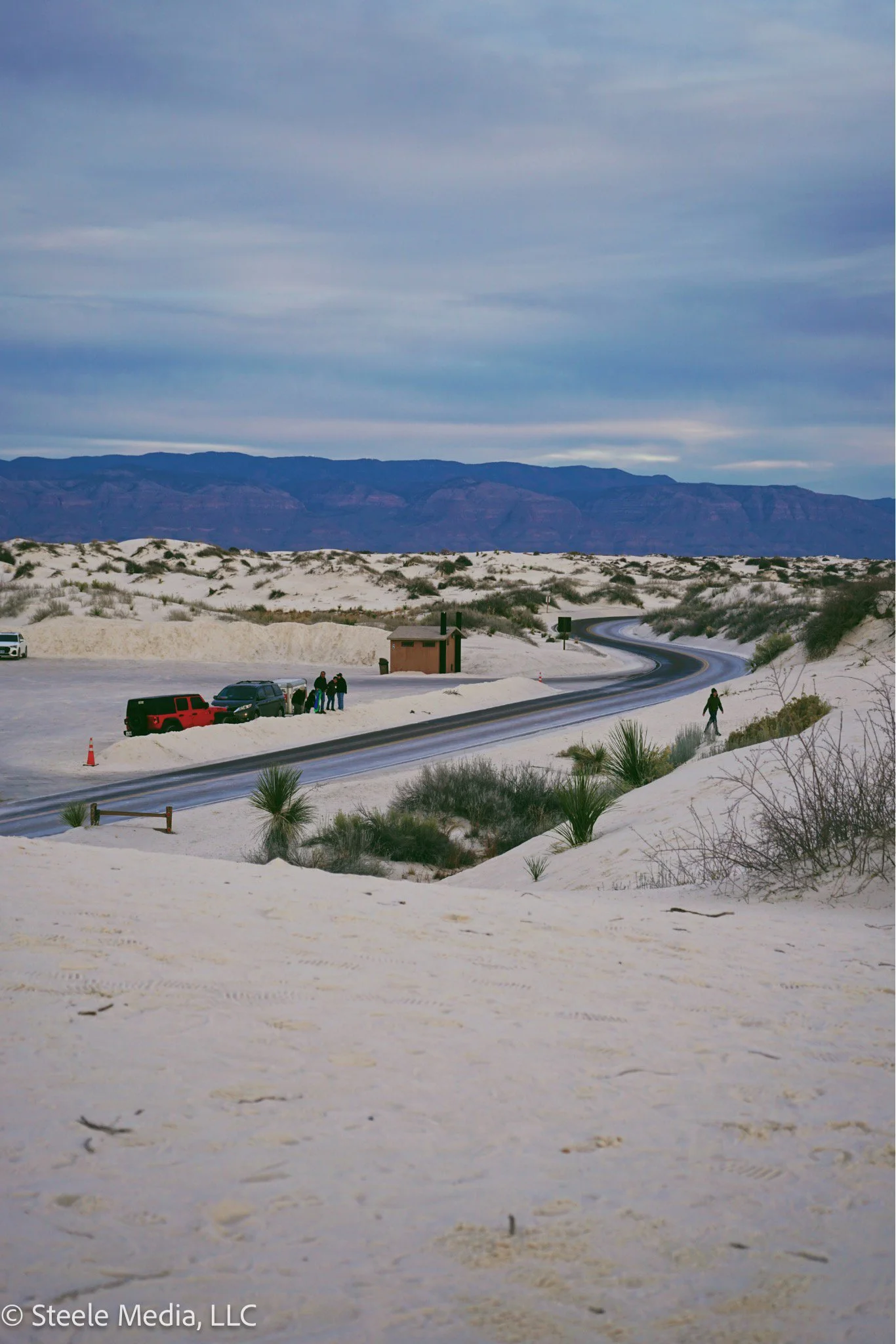 Snow-covered desert landscape with a winding road, small building, vehicles, and people, with mountains in the background and cloudy sky above.