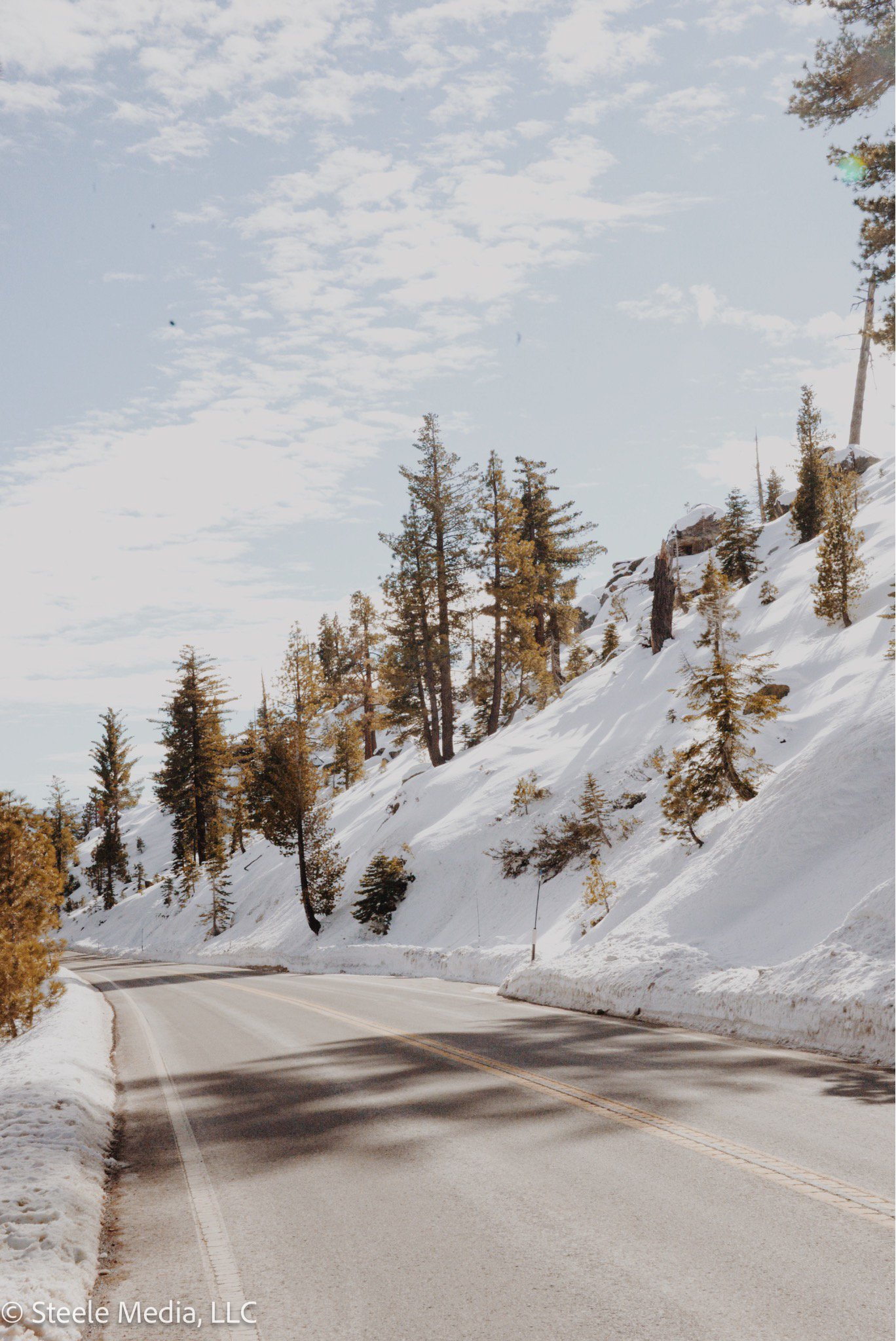 Snow-covered mountain road with trees on a slope, under a partly cloudy sky.