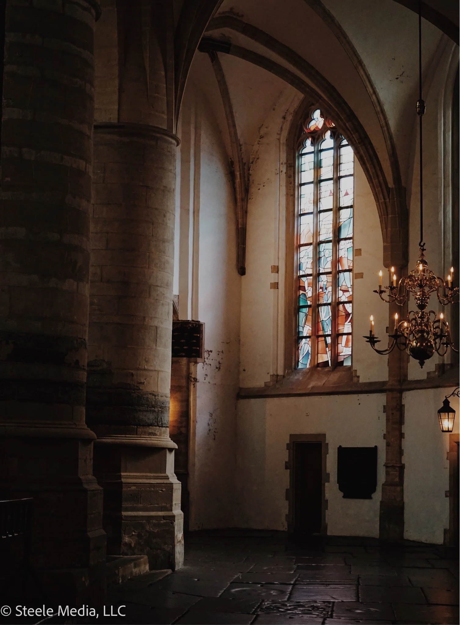 Interior of a historic church with stained glass windows, stone columns, a chandelier, and hanging lamps.