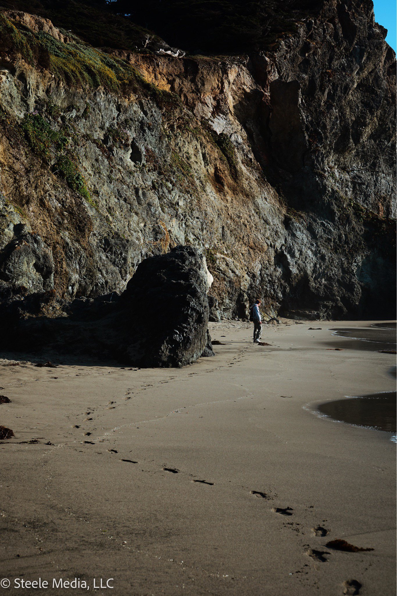 A person walking along a sandy beach near tall, rugged cliffs with green vegetation and large dark rocks, under a clear blue sky.