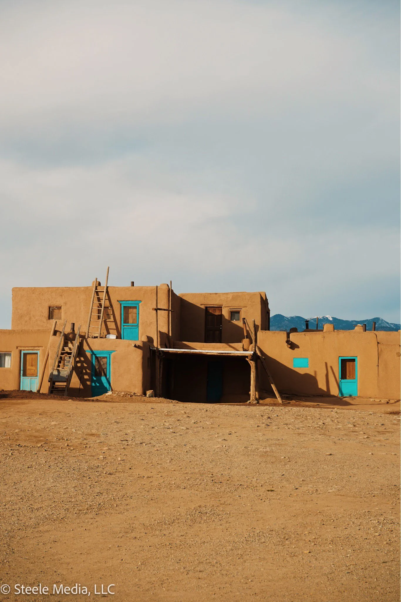 A traditional adobe-style building with blue door and window accents in a barren desert landscape with mountains in the background.