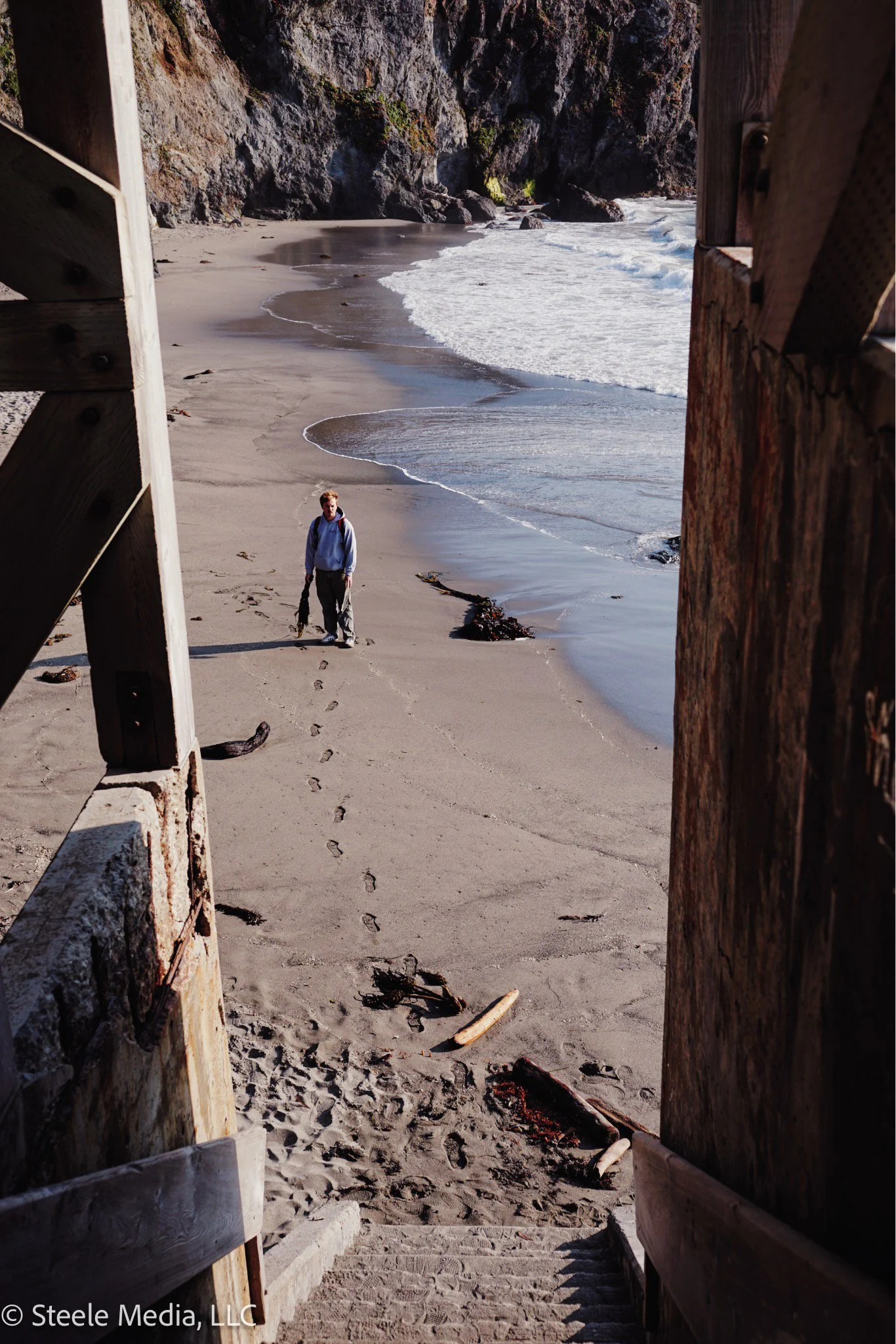 A person walking on a sandy beach viewed from a staircase entrance with wooden railings, with ocean waves and rocky cliffs in the background.