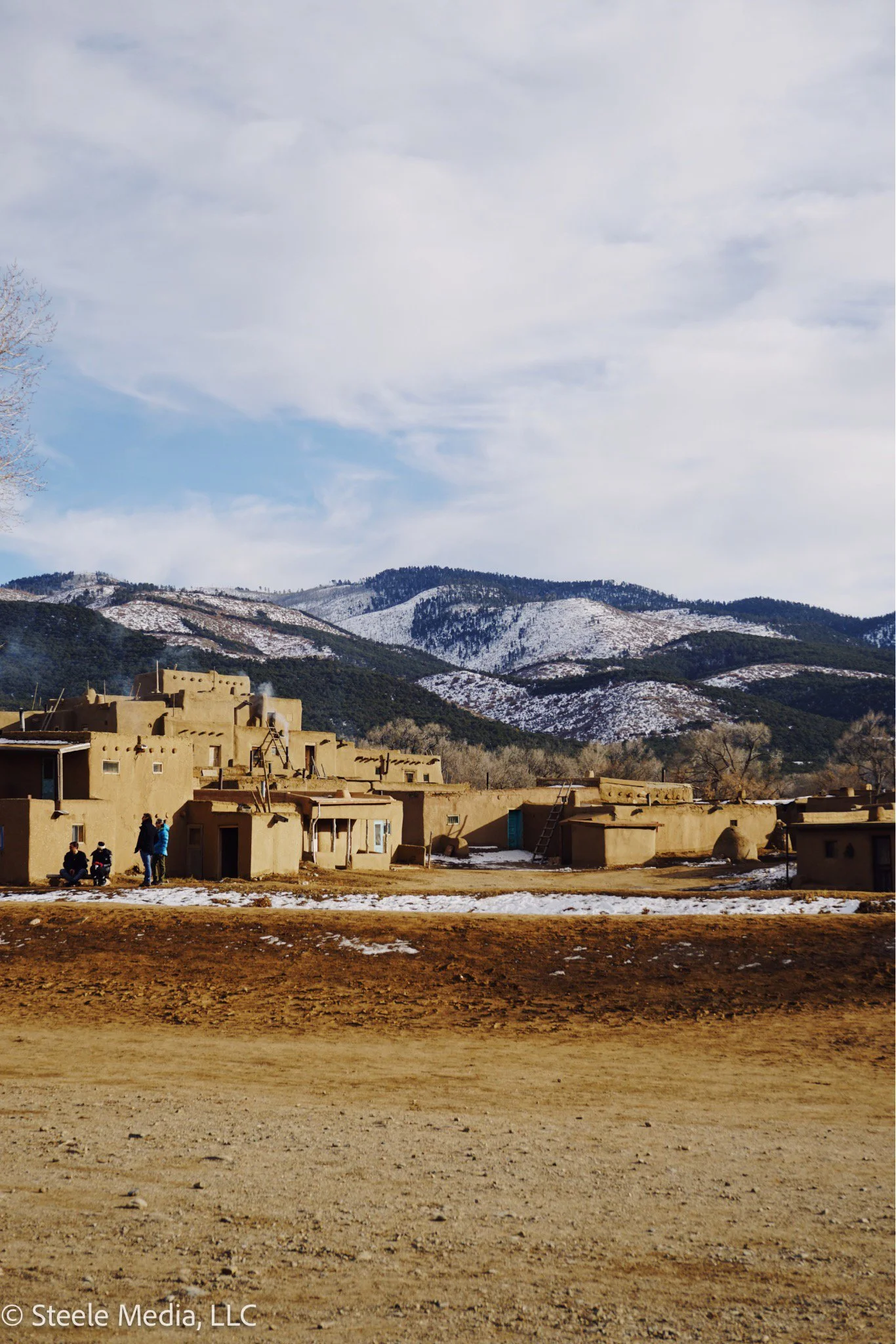 A rural adobe village with traditional flat-roofed buildings, some with ladders, set against a backdrop of snow-capped mountains and partly cloudy sky.
