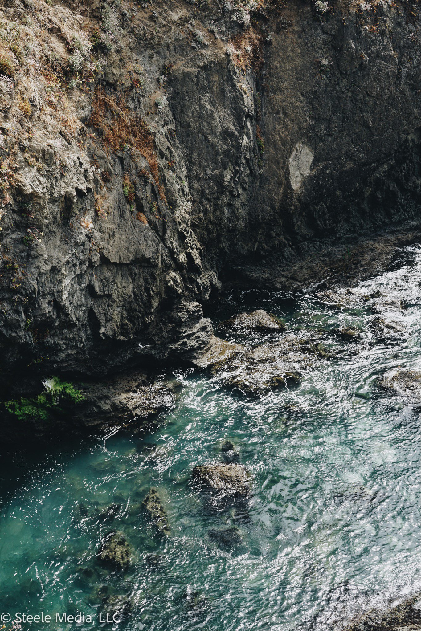 Rocky cliffside along a body of water with white foam and algae-covered rocks in the water.