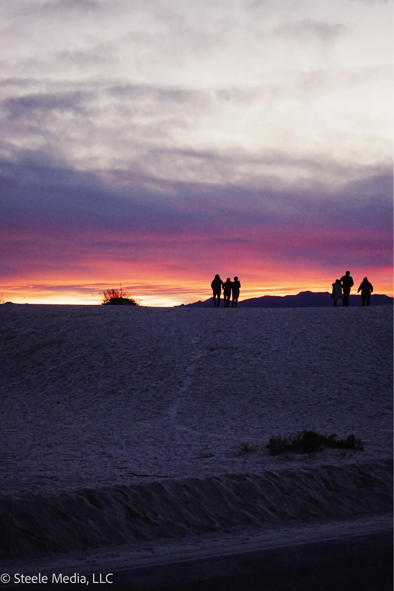 Silhouettes of people walking on a sandy beach during a colorful sunset with purple, pink, orange hues and distant mountains.