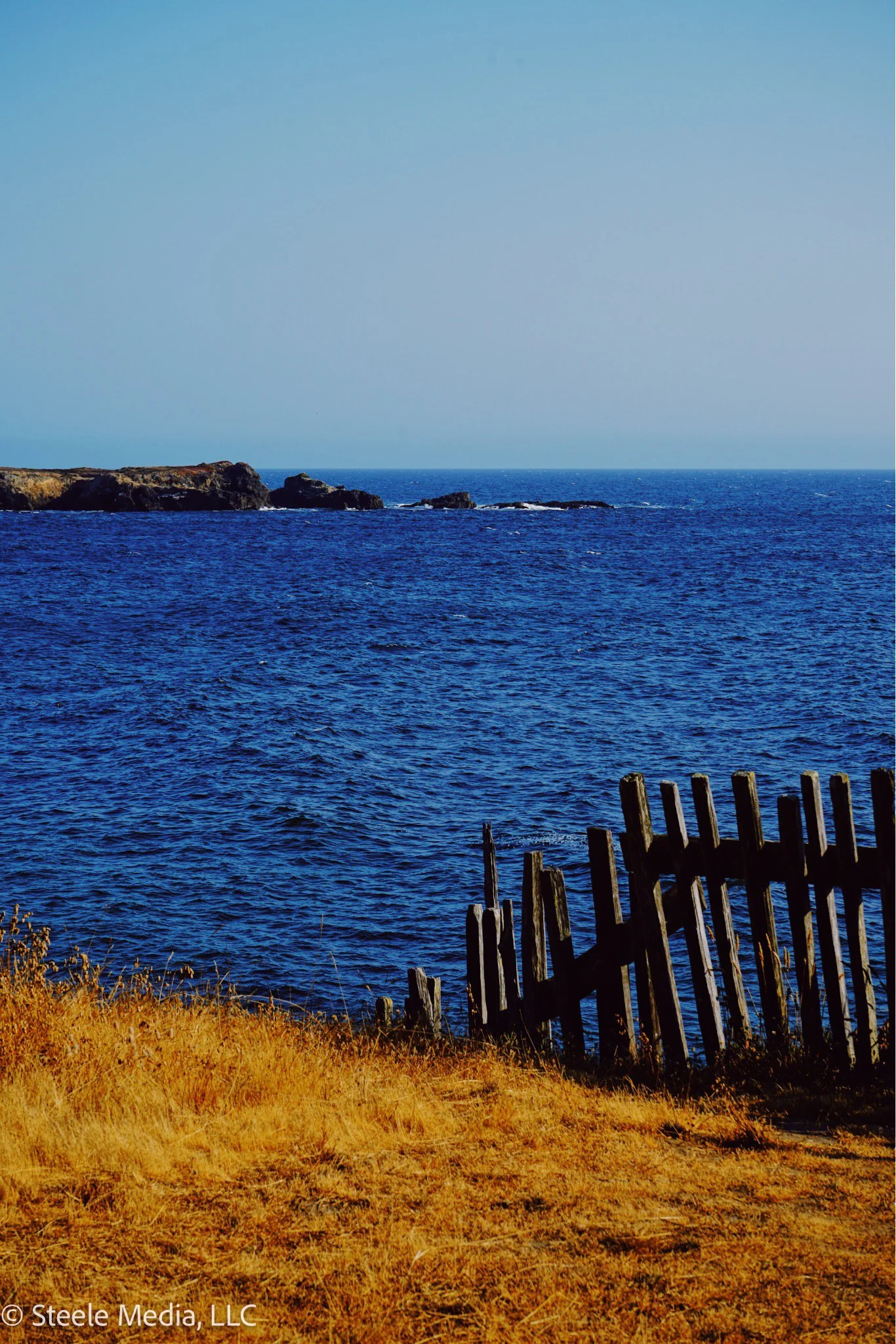 Ocean view with blue water and rocky shoreline, a hay-covered foreground, and a wooden fence in the lower right.
