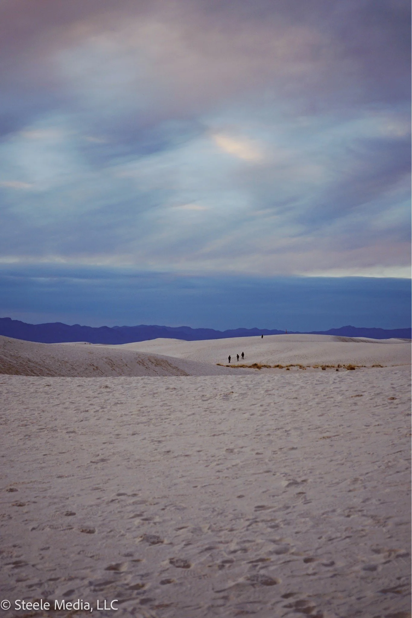 Desert landscape with rolling sand dunes, a cloudy sky, and a few people walking in the distance.