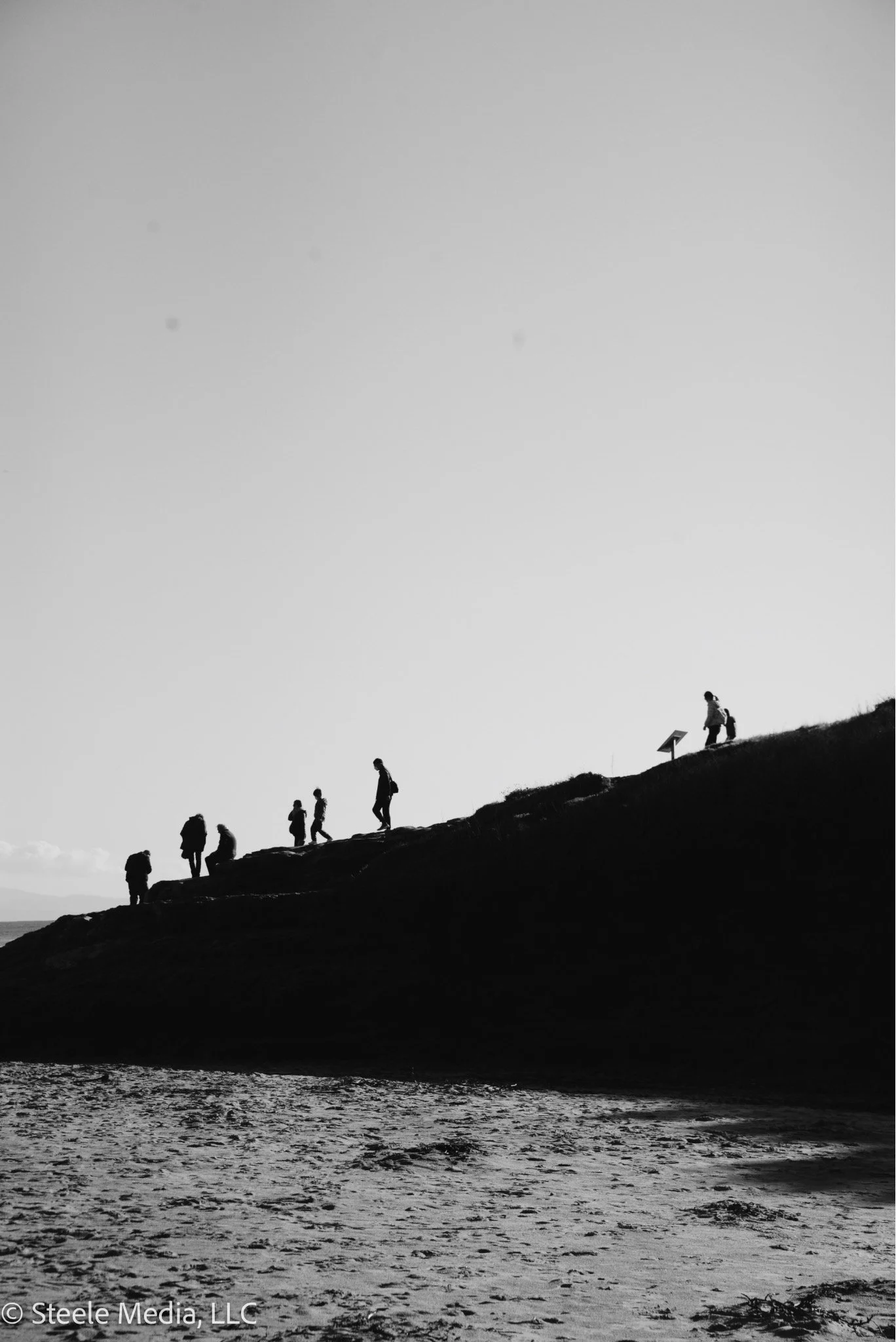 Silhouettes of seven people hiking up a hillside, some with backpacks, towards a sign on the top right, with a mostly clear sky in the background and a sandy area at the bottom.