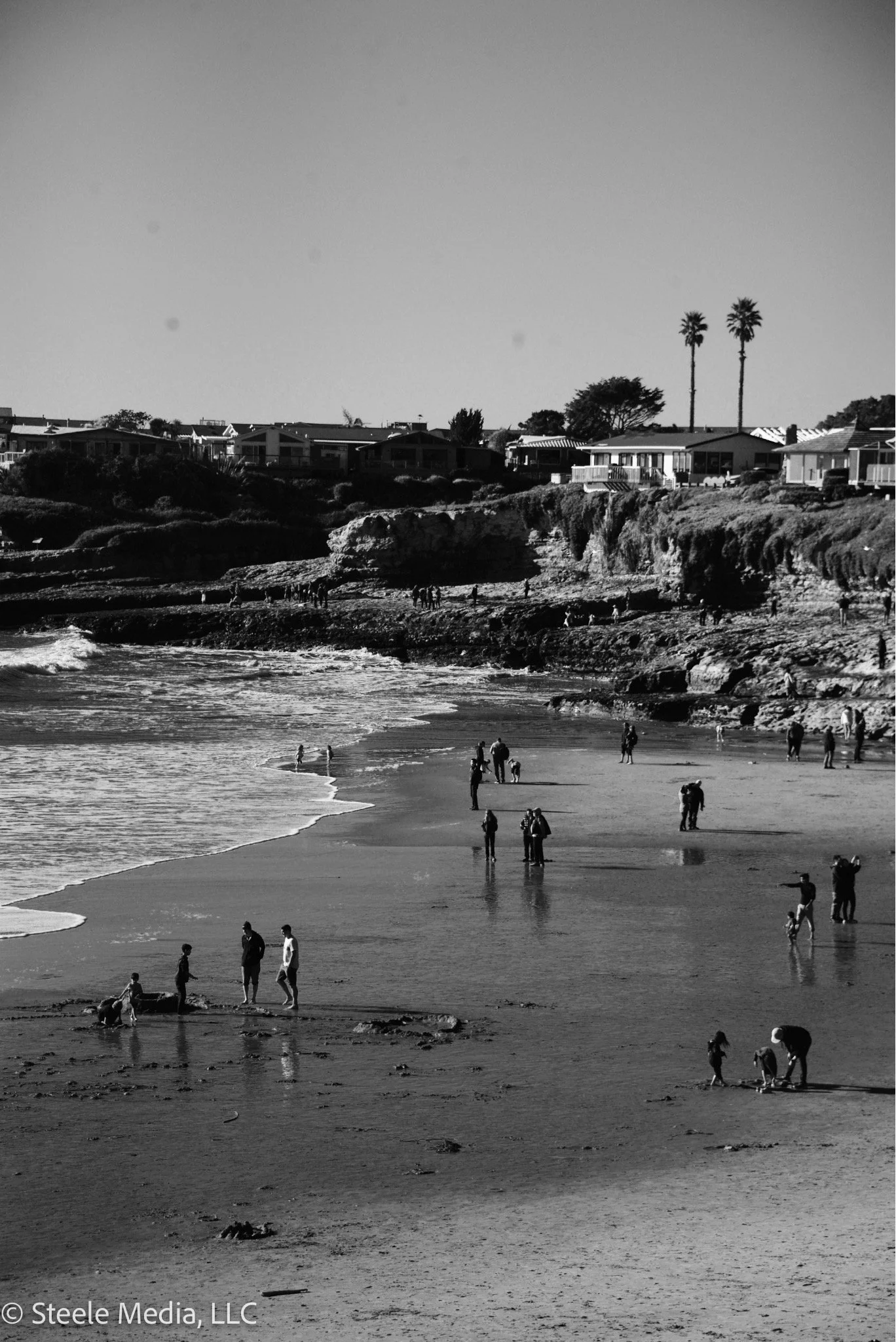 People walking and playing on a beach with cliffs and residential houses in the background.