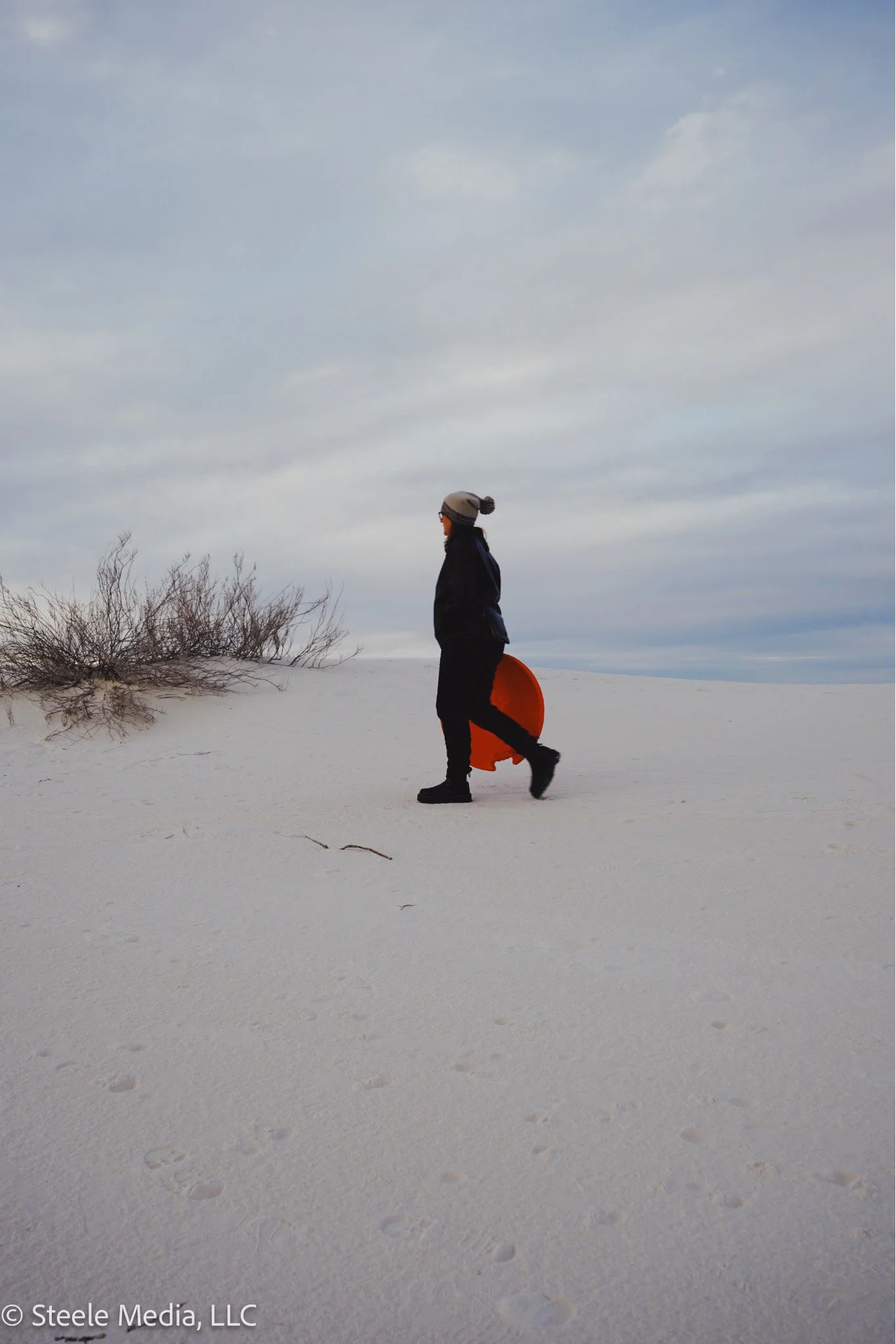 A person walking in a snowy landscape, wearing a dark jacket, pants, and a knit hat with a pom-pom, carrying an orange sled.