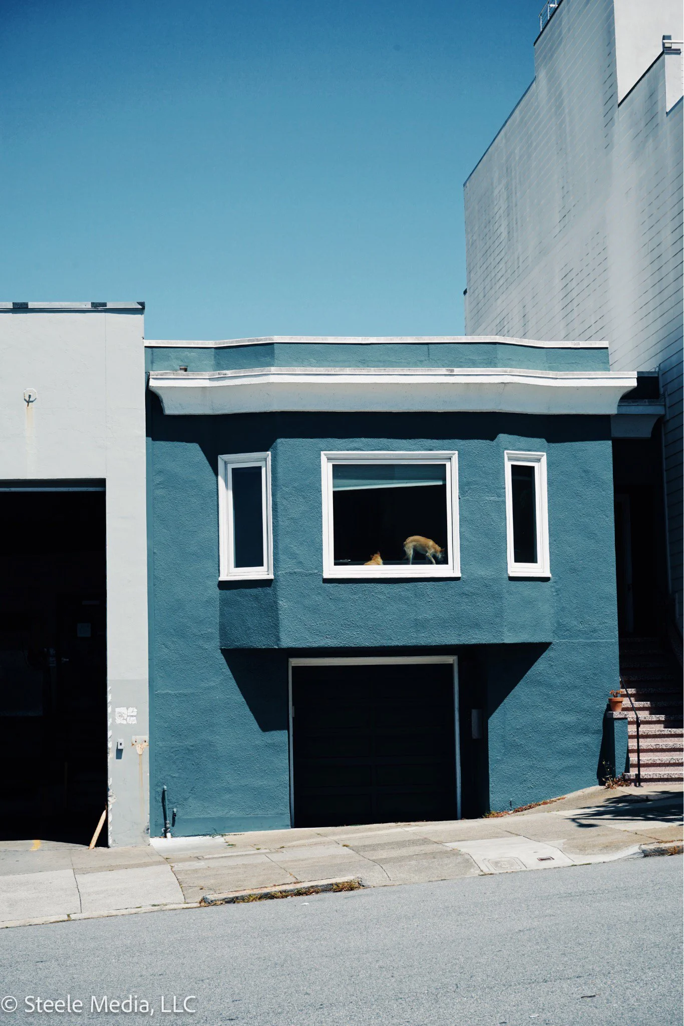A blue two-story house with a garage on the ground level and a window on the upper level. Two cats are visible through the upper window, one inside the house and one on the window ledge. Surrounding buildings are white, and there are stairs on the ri