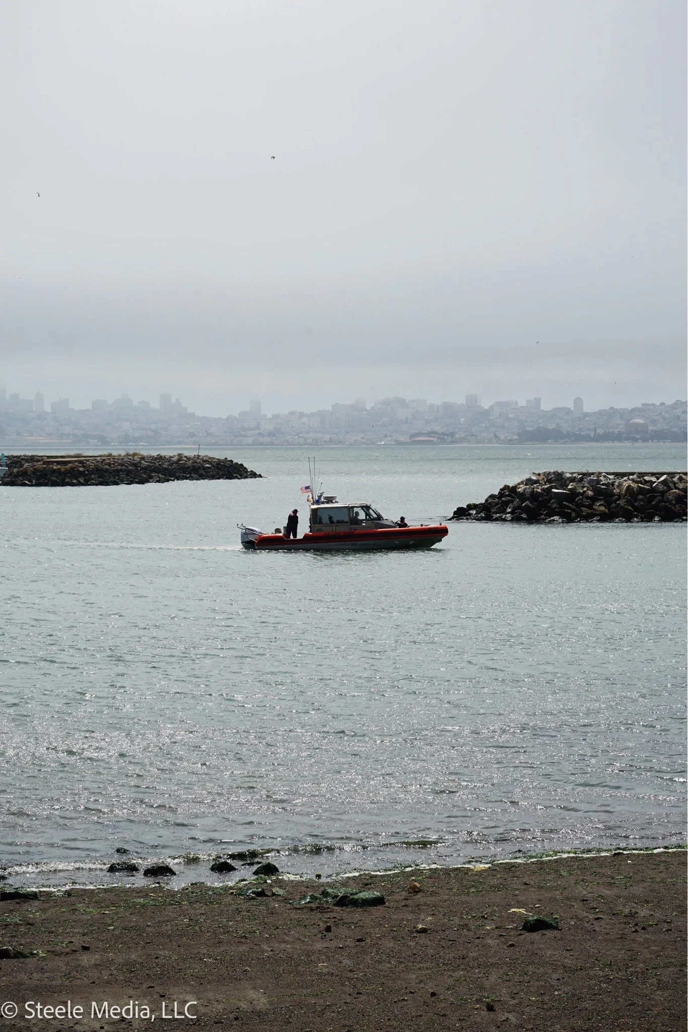 A boat with a small cabin and two people on board, sailing in a harbor near rocky breakwaters with a city skyline in the background under cloudy sky.