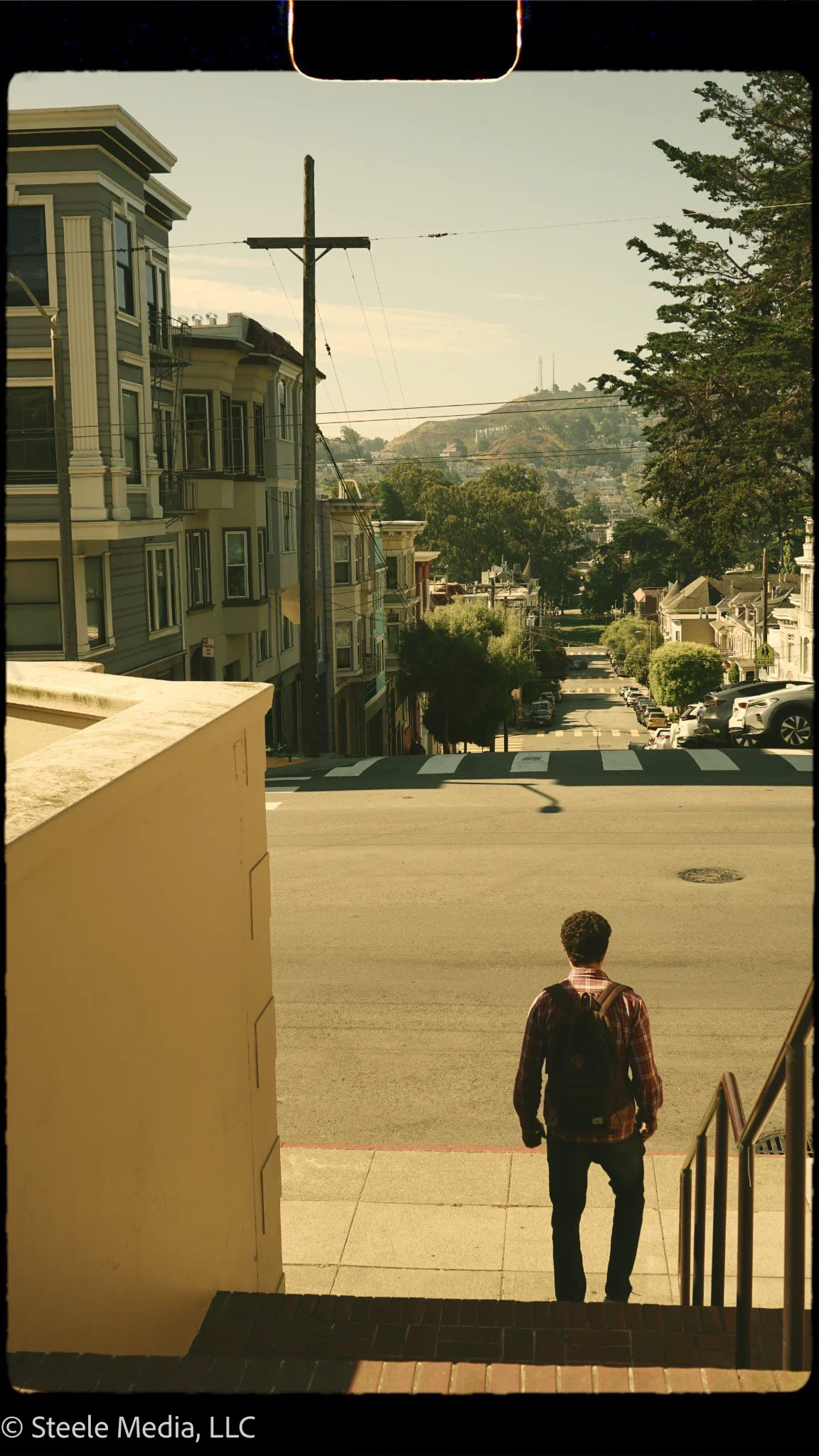 A person with a backpack walking down the steps onto a city street sidewalk, with residential buildings on the left and parked cars on the right, under a partly cloudy sky.