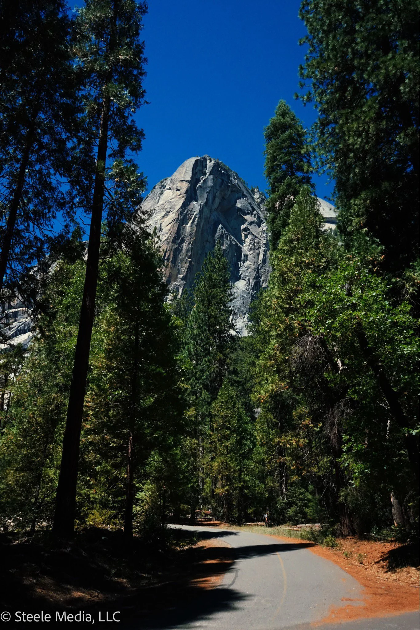 A mountain view with tall, green trees along a winding road and a large granite mountain in the background under a clear blue sky.
