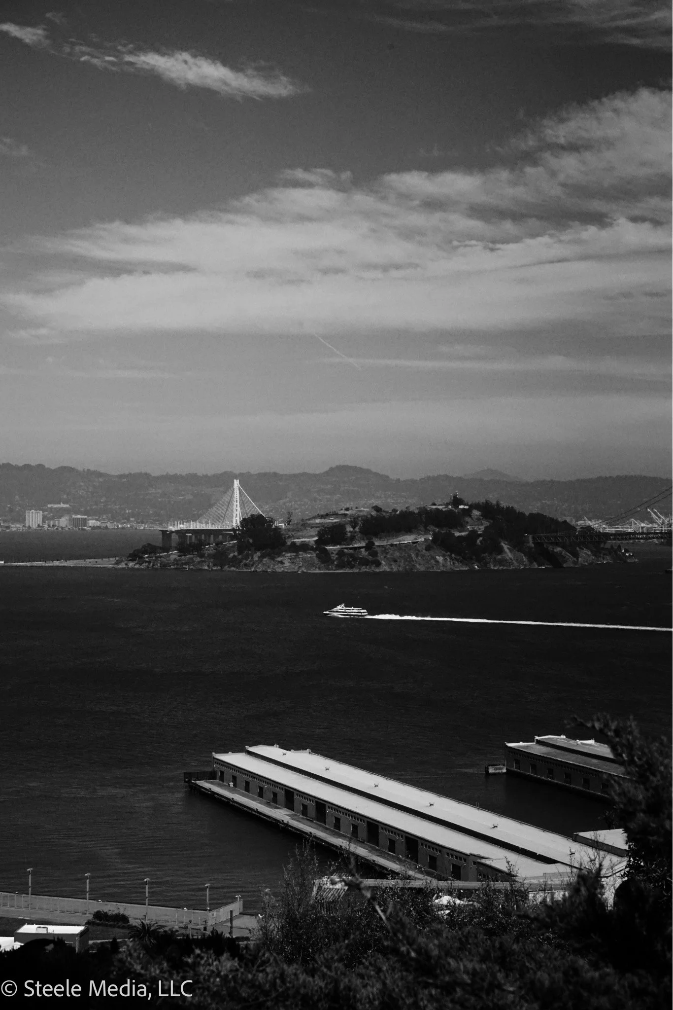 Black and white photo of San Francisco Bay with Alcatraz Island, a bridge, and a yacht creating a wake in the water.