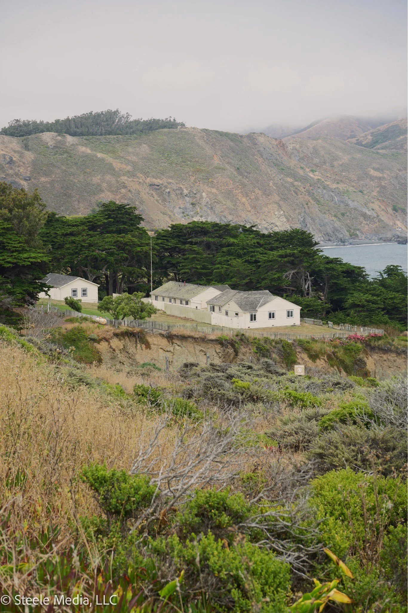 A coastal scene with a few houses, trees, and hills in the background. The sky is overcast, and the vegetation in the foreground is dry and green.
