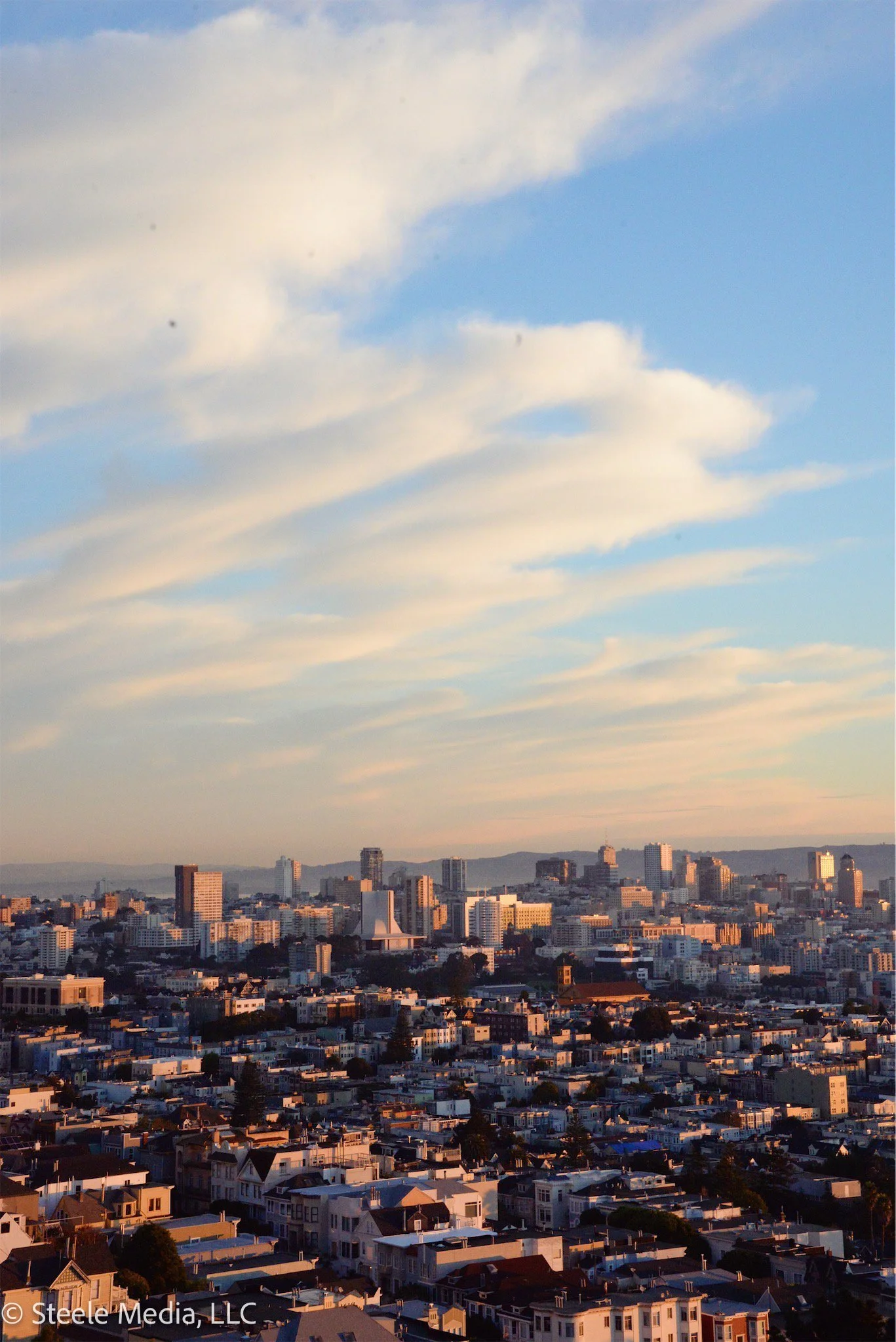 A city skyline at sunset with a blue sky and wispy clouds, featuring various tall buildings and residential neighborhoods in the foreground.