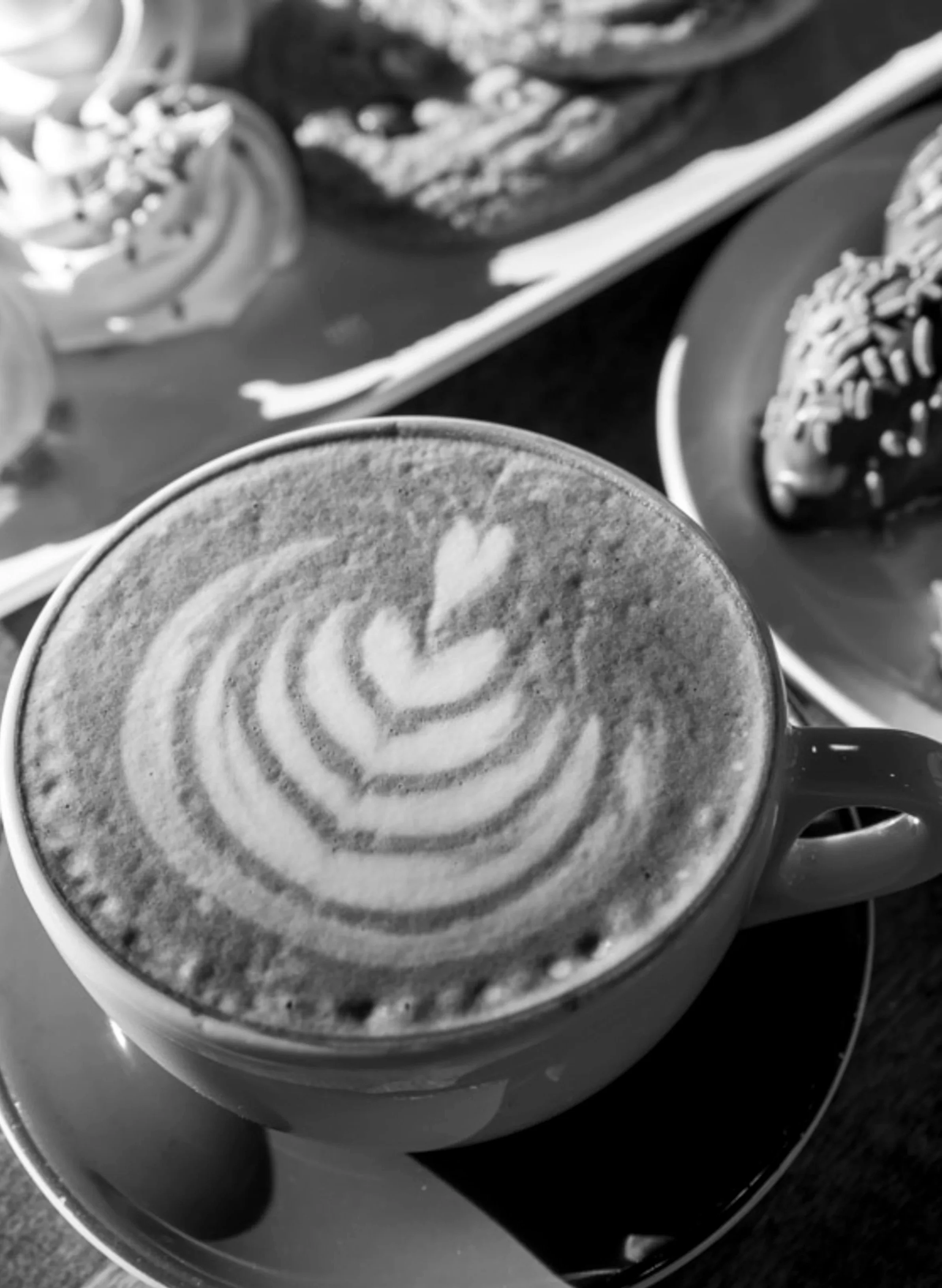 Close-up of a coffee cup with latte art foam design, with various donuts in the background on plates and a tray.