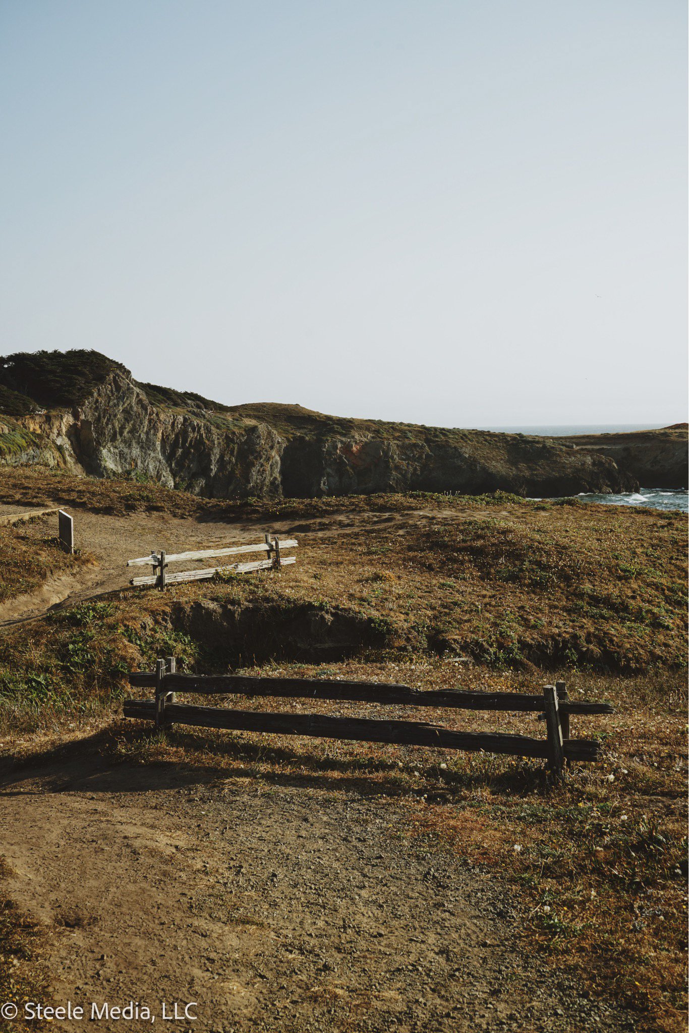 A scenic coastal landscape with dirt paths, wooden fences, grassy terrain, rocky cliffs, and the ocean in the distance under a clear sky.